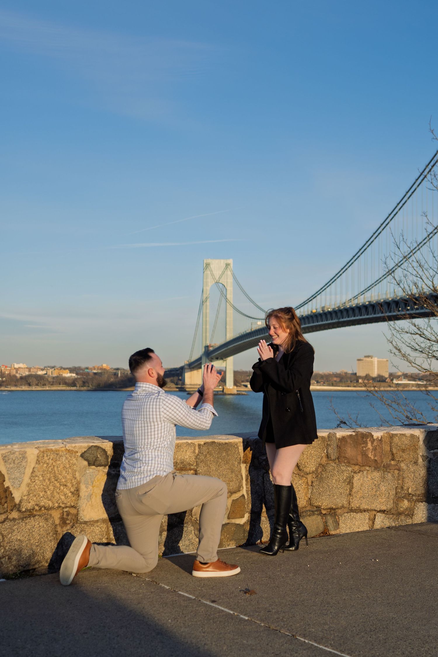 Man kneeling on one knee proposing to woman by the Verrazzano-Narrows Bridge at golden hour.