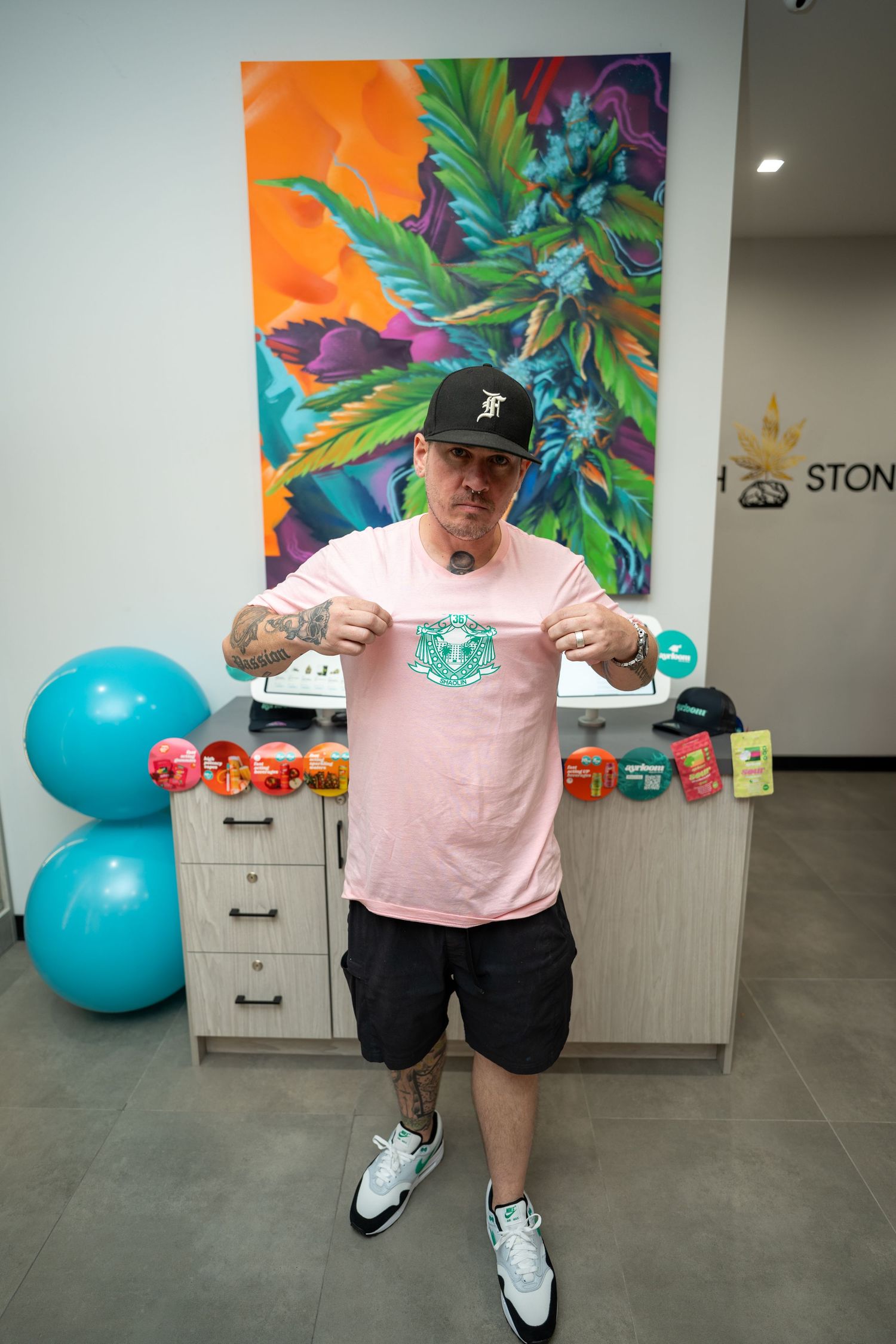 Tattooed man in pink shirt pointing to logo tee inside a cannabis dispensary.