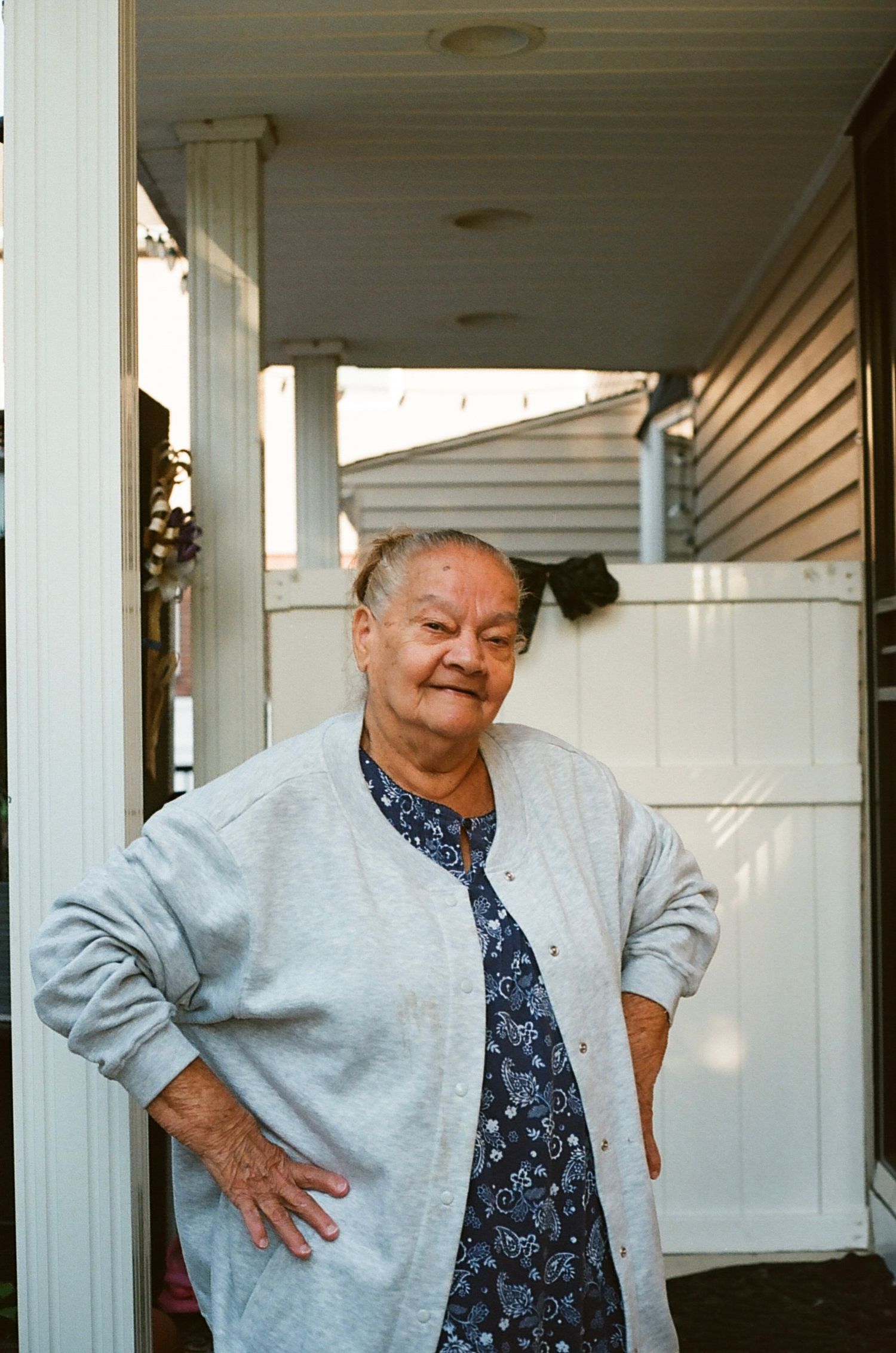 Elderly woman in gray cardigan and floral shirt standing on porch with hands on hips smiling.