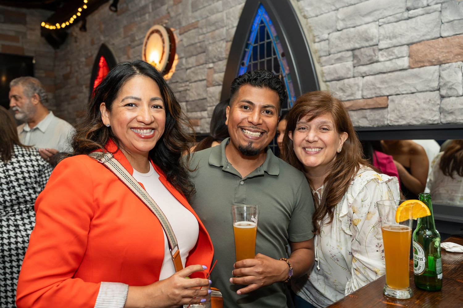 Three people smiling and holding drinks at a lively indoor bar event, one woman wearing devil horns.