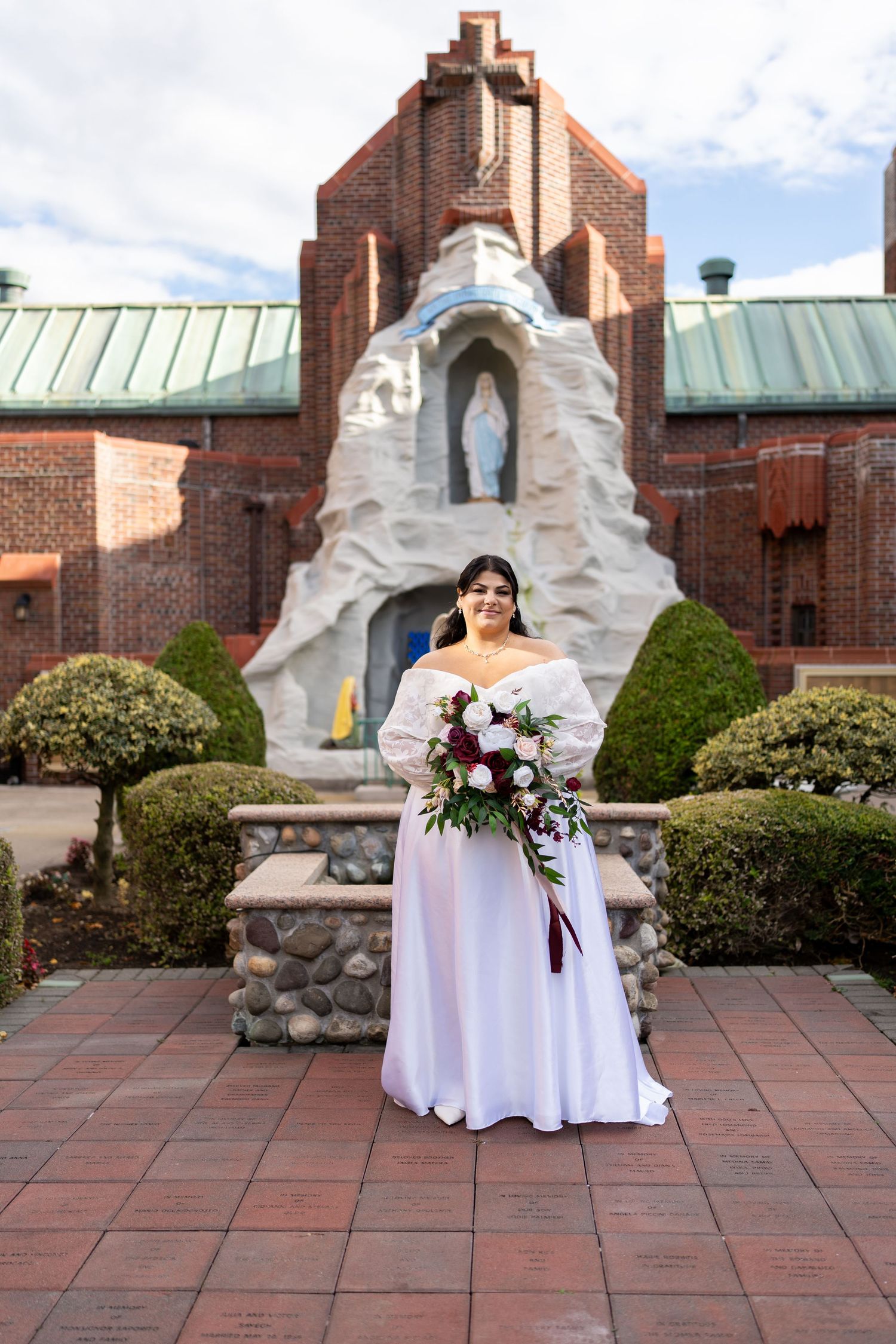 Bride in off-shoulder white gown holding burgundy and white bouquet standing before a brick church grotto statue.