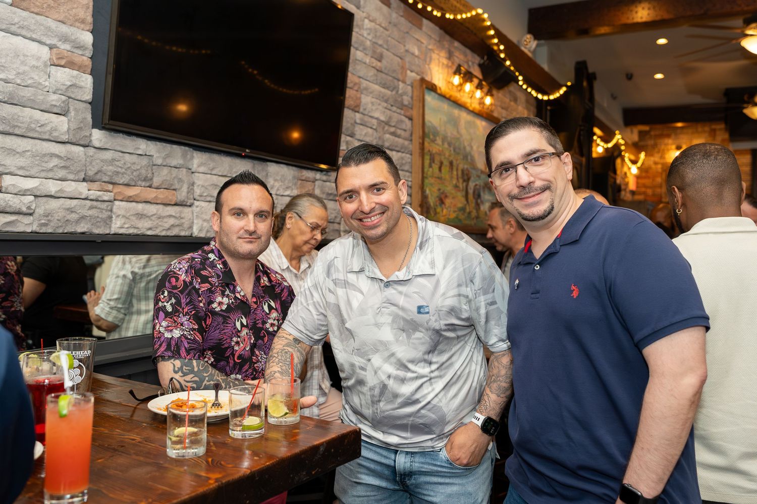 Three men smiling together at a rustic restaurant bar with cocktails and appetizers on the table.