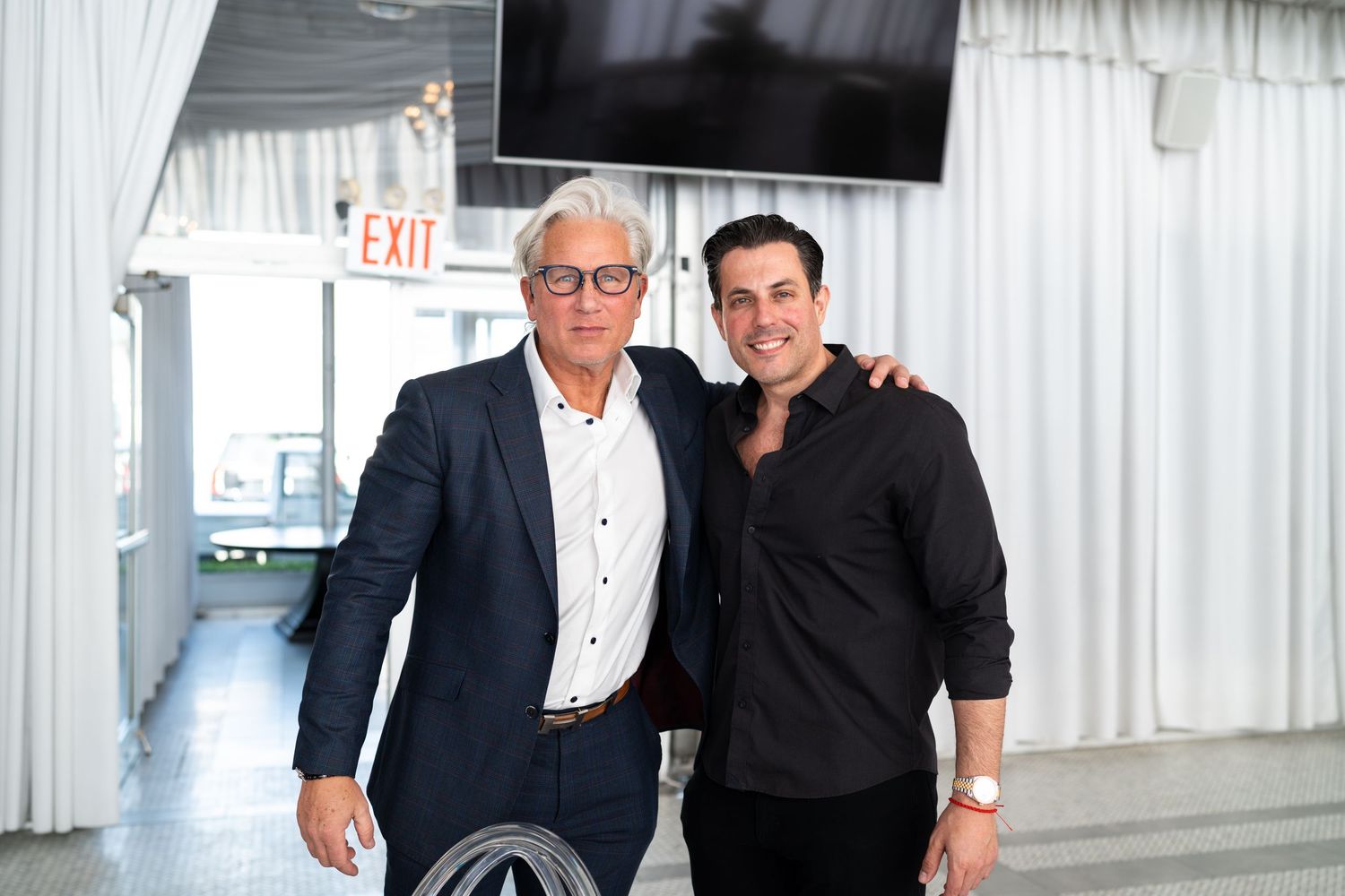 Two men in business casual attire smiling together at an indoor event venue with white draping.