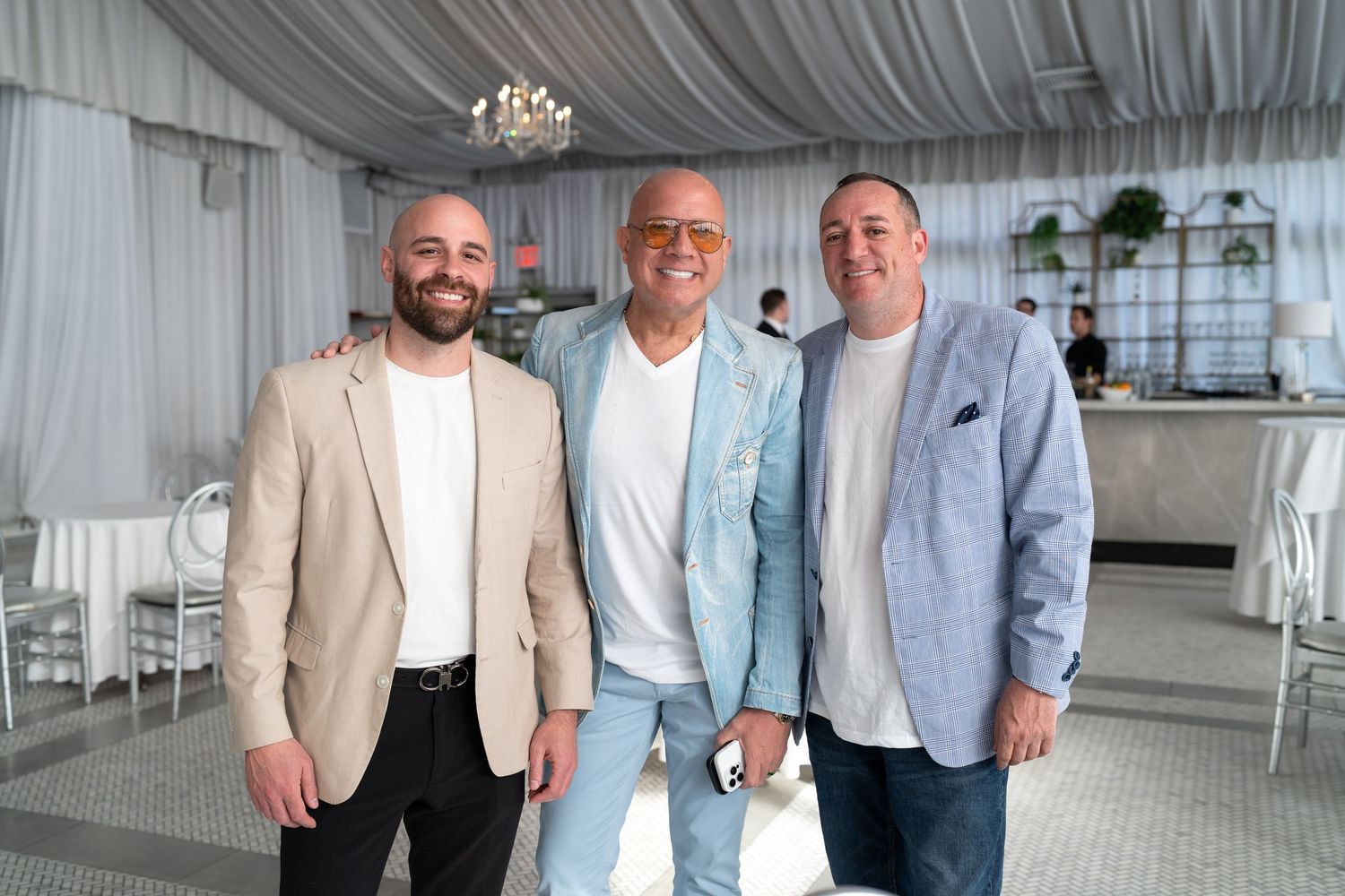 Three smiling men in smart casual blazers pose together inside a white draped event tent venue.