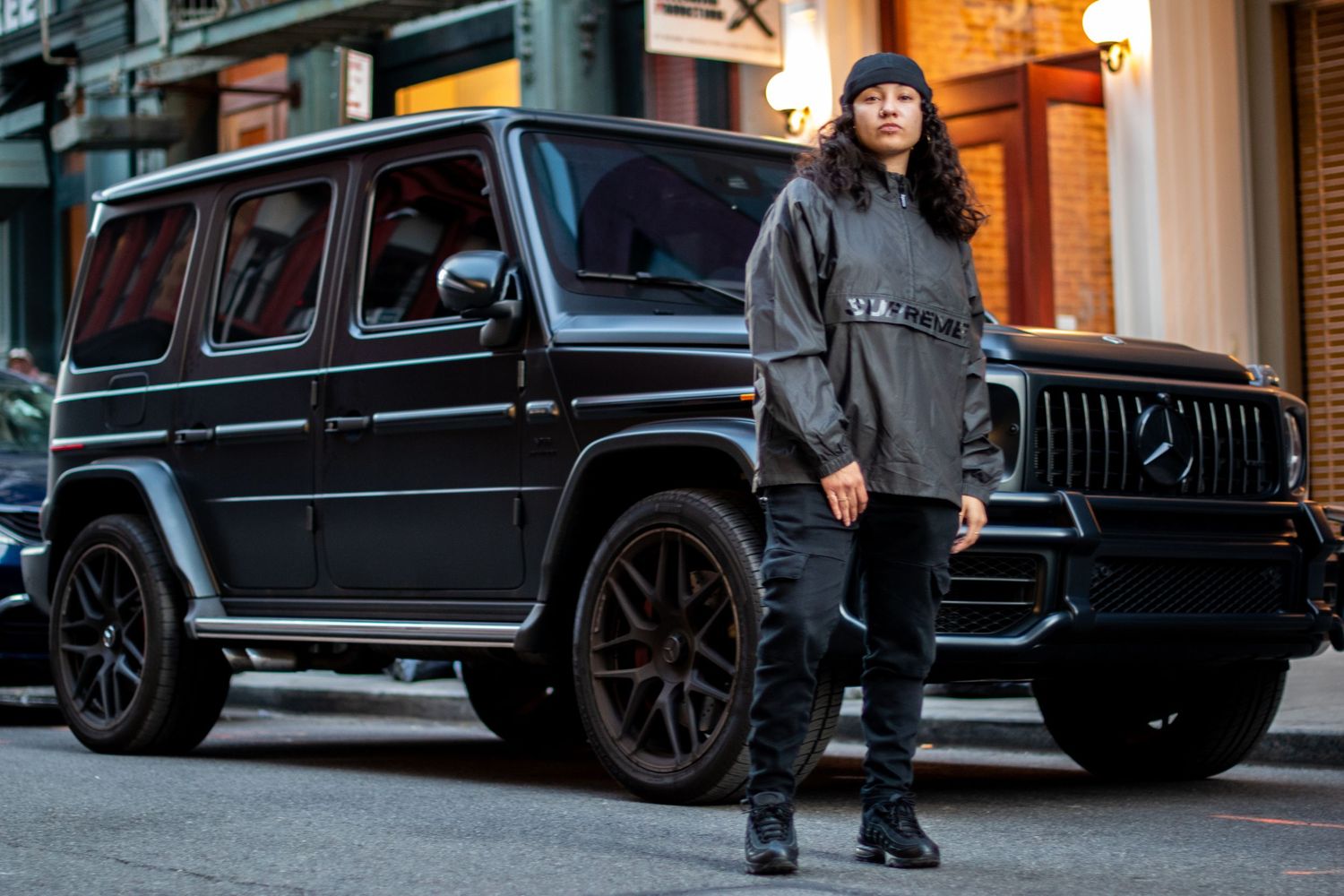 A woman in streetwear stands confidently beside a black Mercedes-AMG G-Class SUV outdoors.