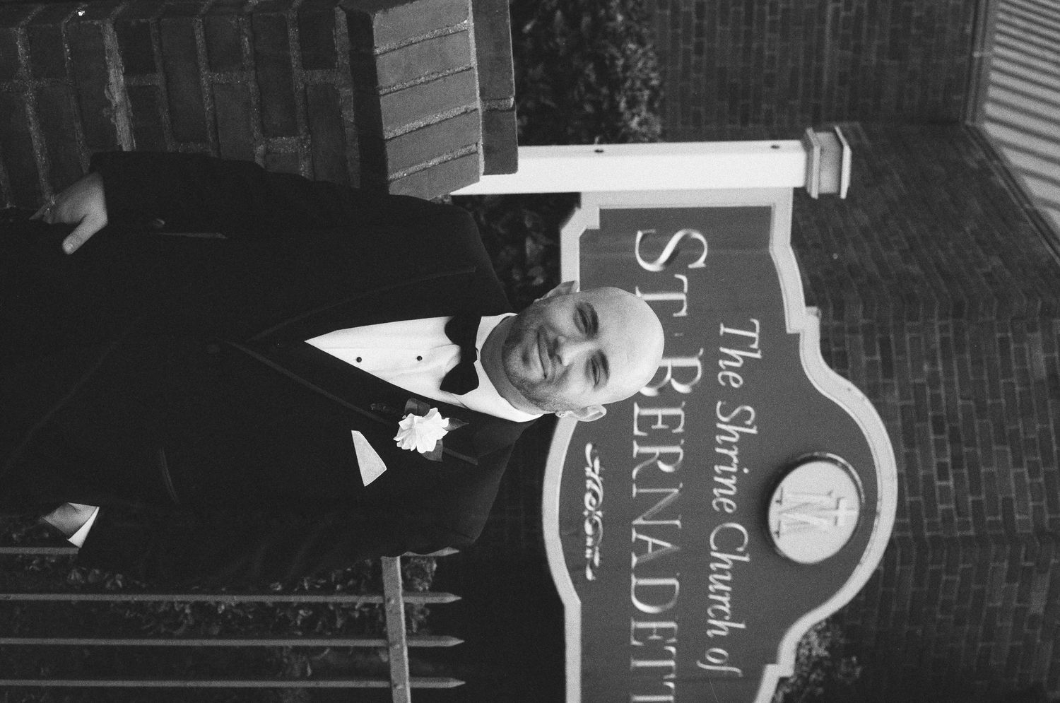 Man in tuxedo posing beside The Shrine Church of St. Bernadette sign in black and white photo.
