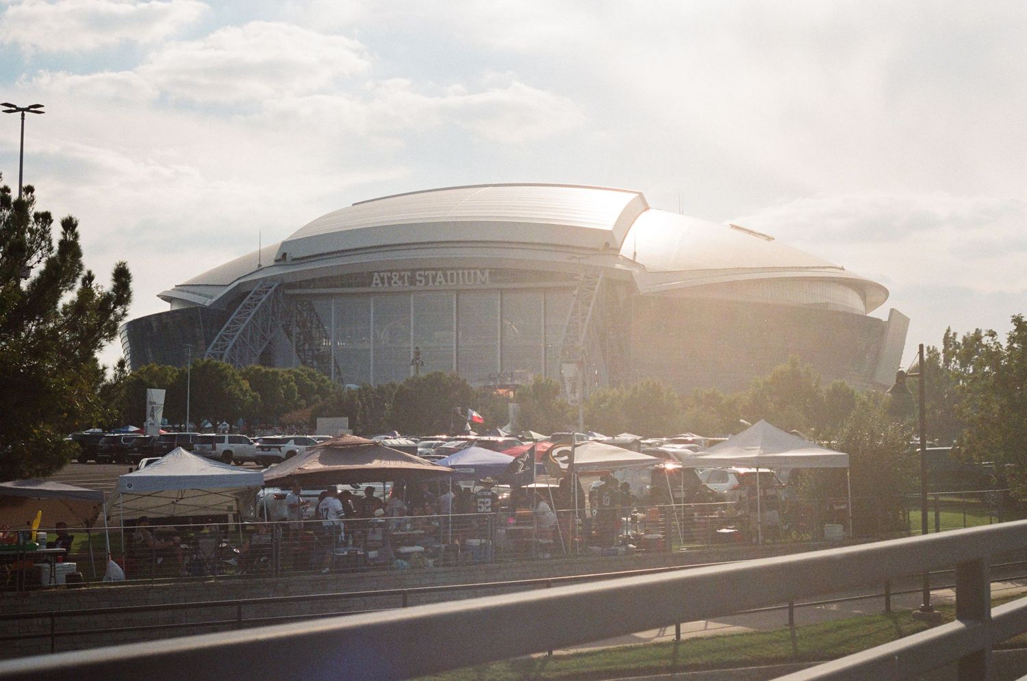 Aerial view of a large domed stadium with outdoor vendor tents in a sunny parking lot.
