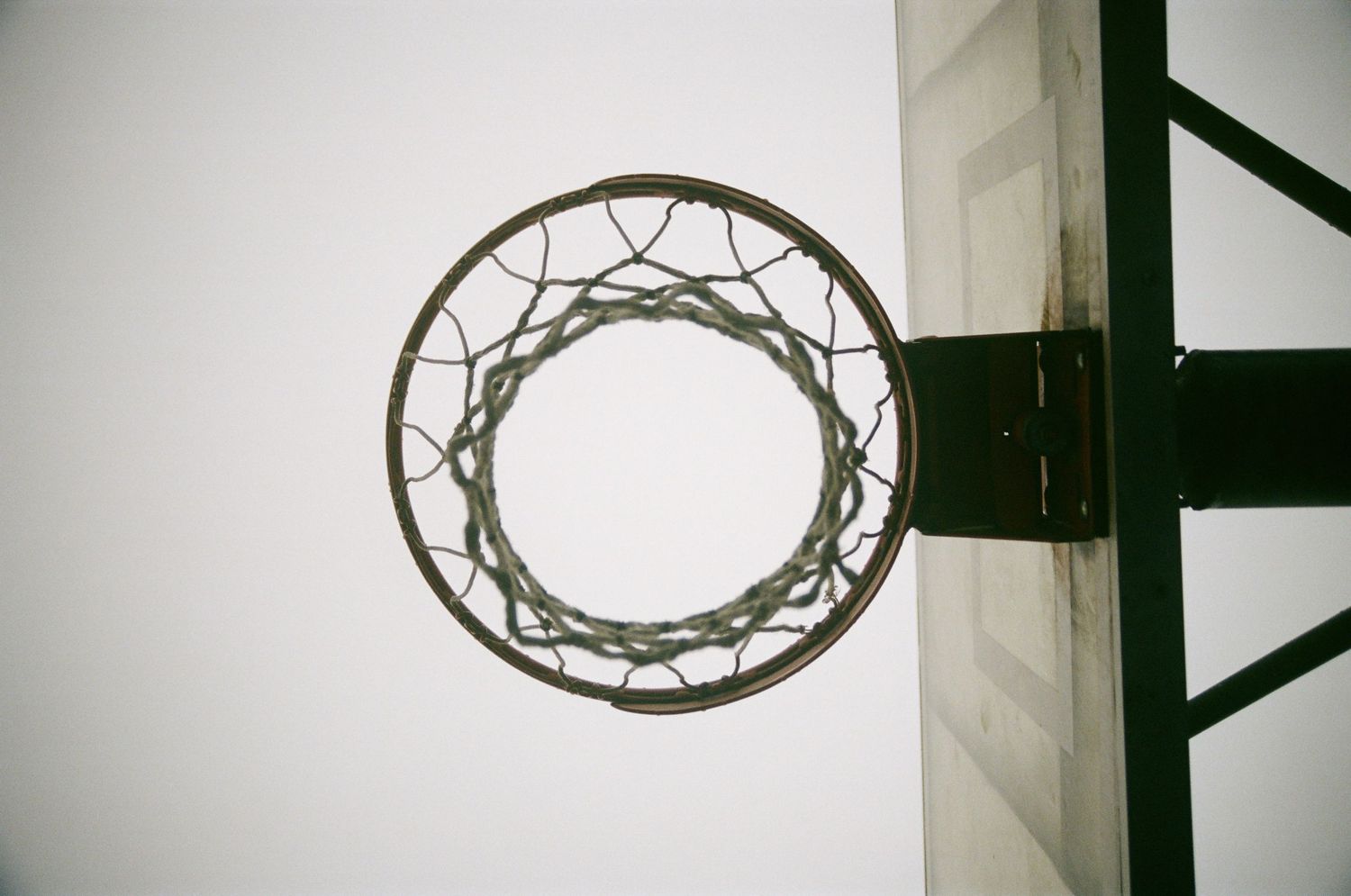 Low angle view looking up through a worn basketball hoop and net against a grey sky.