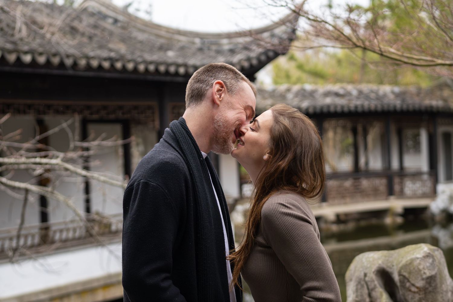 Romantic couple sharing a kiss near a traditional Chinese garden pavilion and pond.