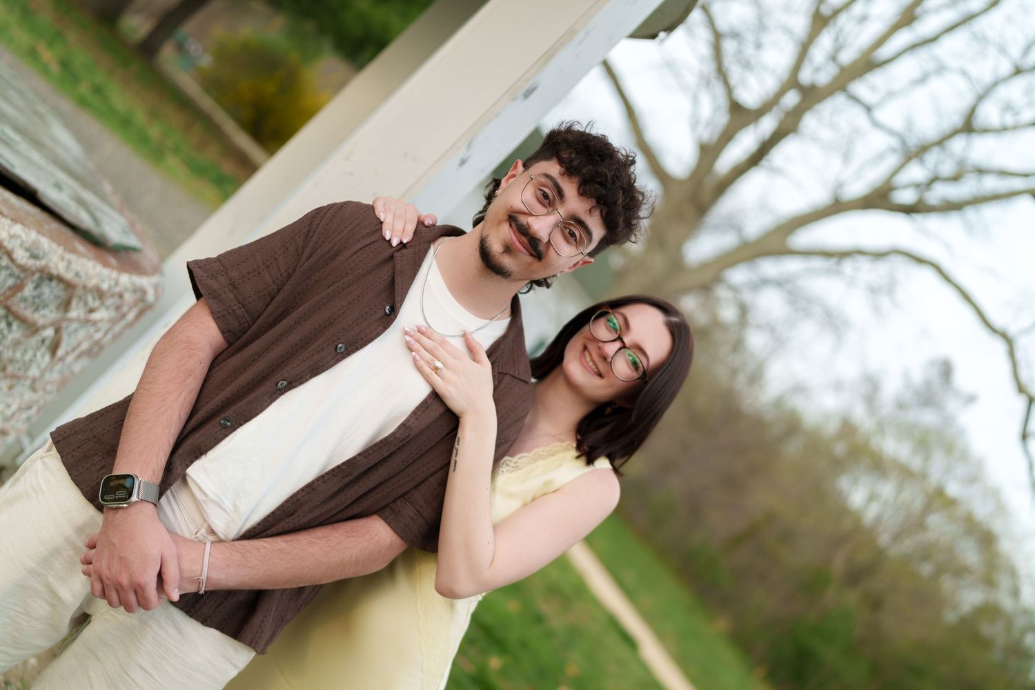 Happy couple embracing and laughing beside a white gazebo pillar in a park with bare trees.