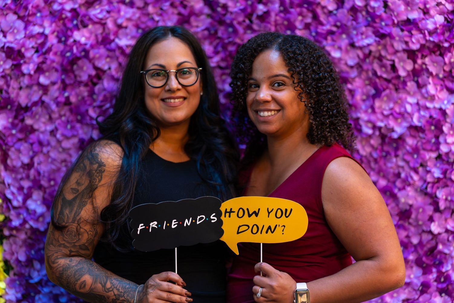 Two women posing with Friends TV show photo booth props at a purple flower wall.