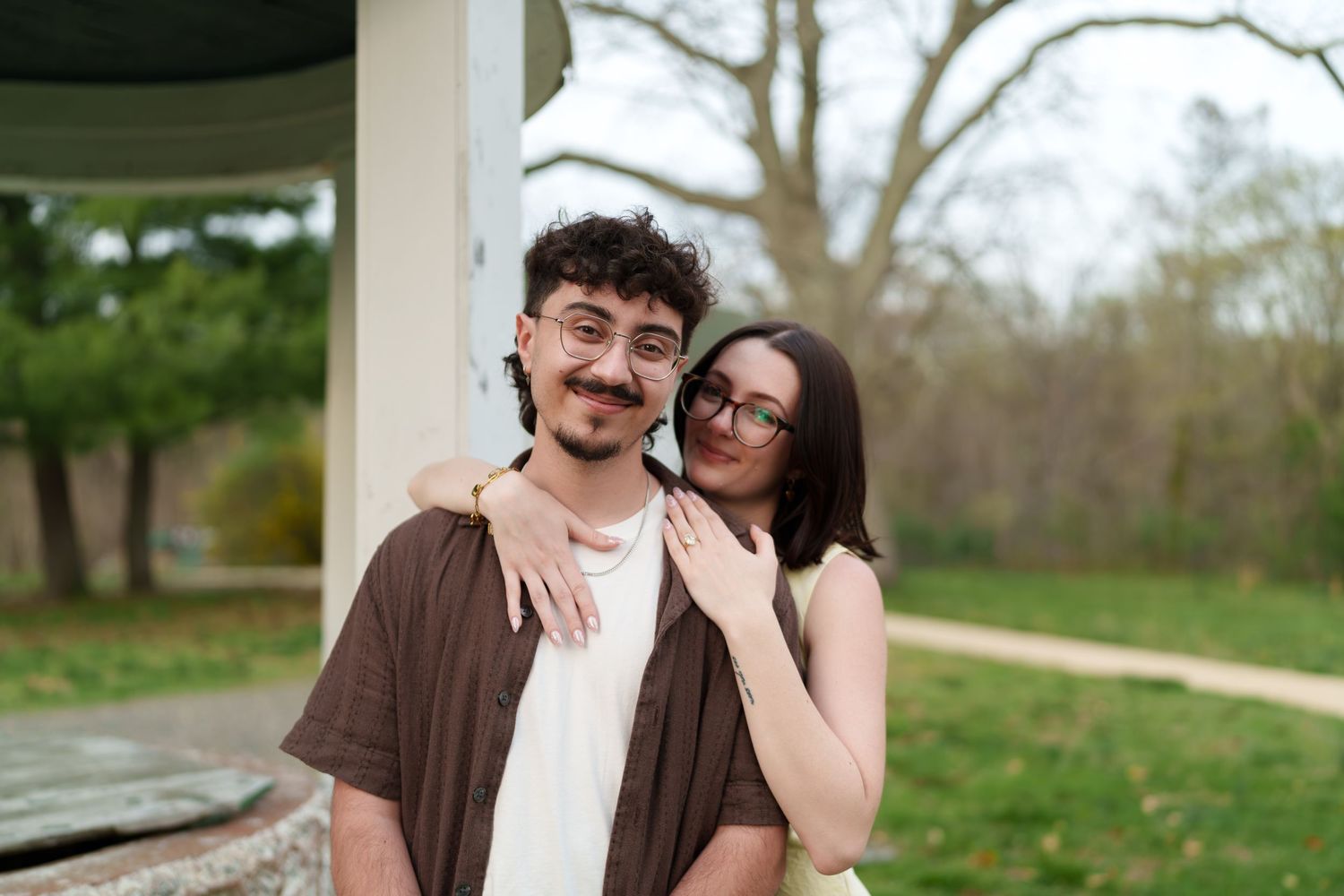 Smiling couple posing together near a white gazebo column in a green outdoor park setting.
