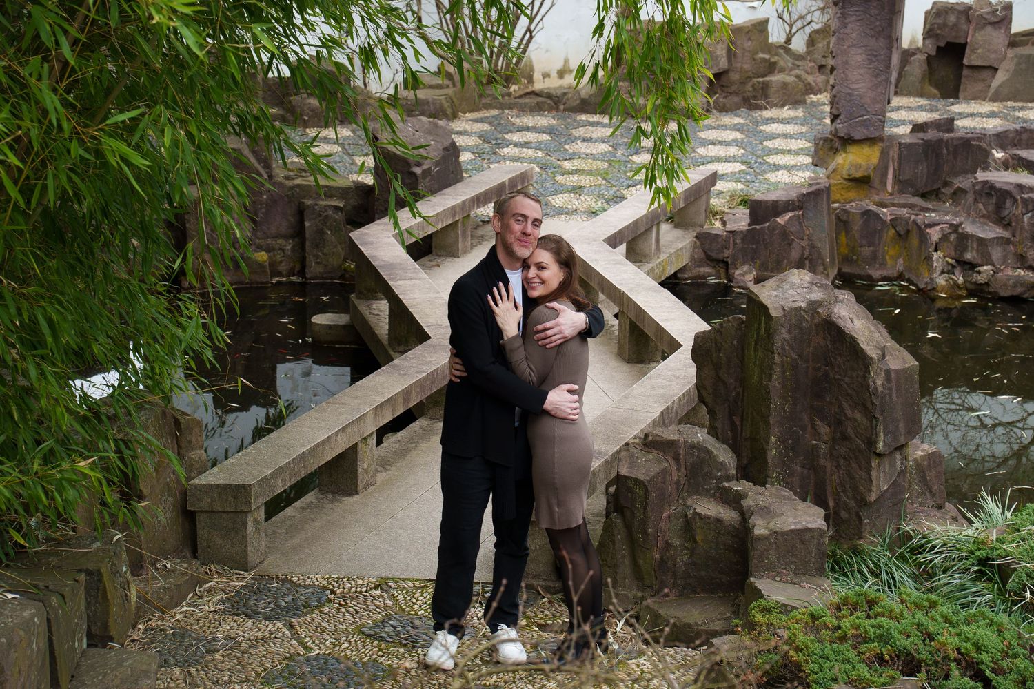 Couple embracing and smiling in a Japanese garden with stone bridge and bamboo.