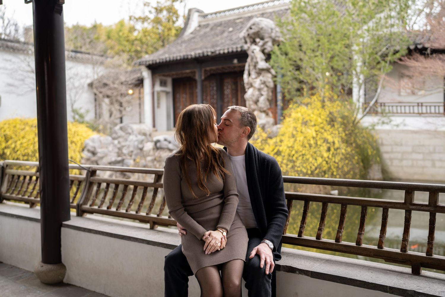 Couple sharing a tender kiss while seated in a scenic Chinese garden with yellow blooms.