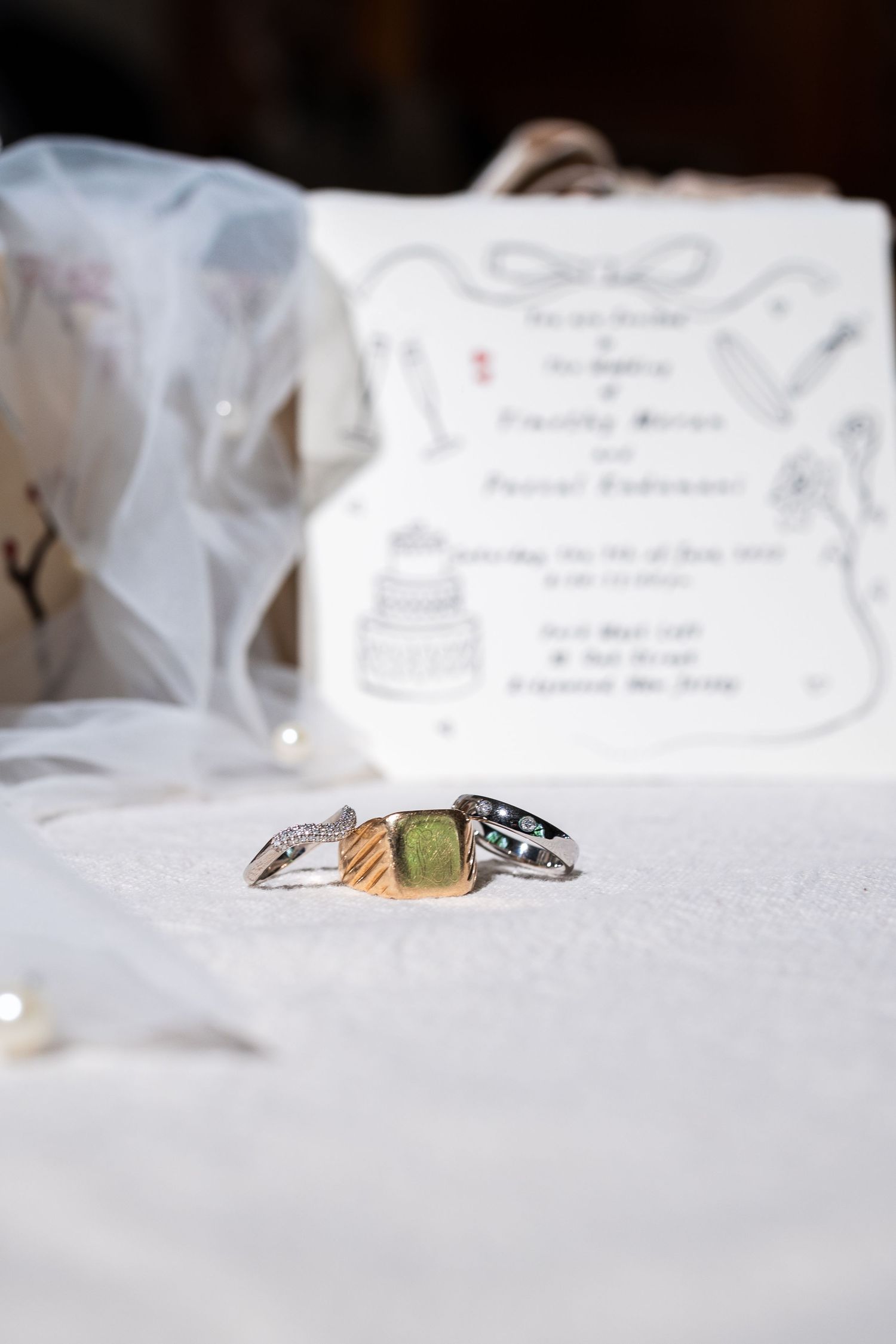 Close-up of wedding rings with green gemstone beside floral wedding invitation and white bridal veil.