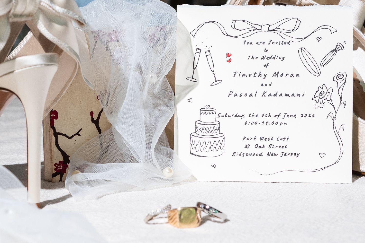 Wedding invitation for Timothy Moran and Pascal Kadamani displayed with bridal shoes, veil, and rings.