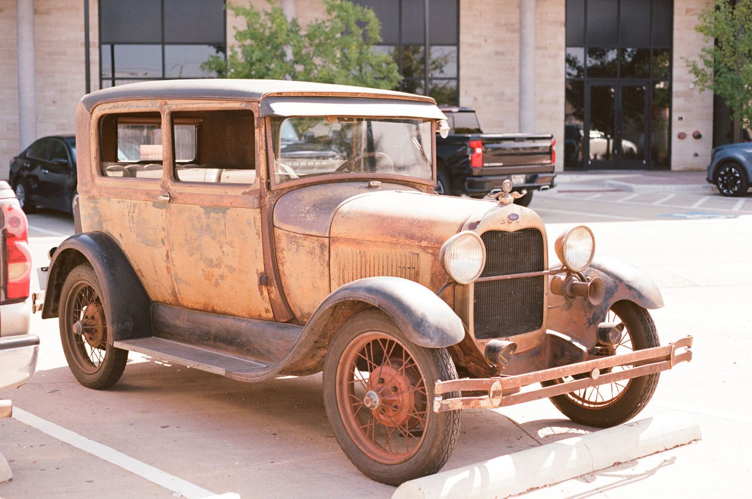 Vintage rusty Ford Model A car parked outdoors in a parking lot on a sunny day.