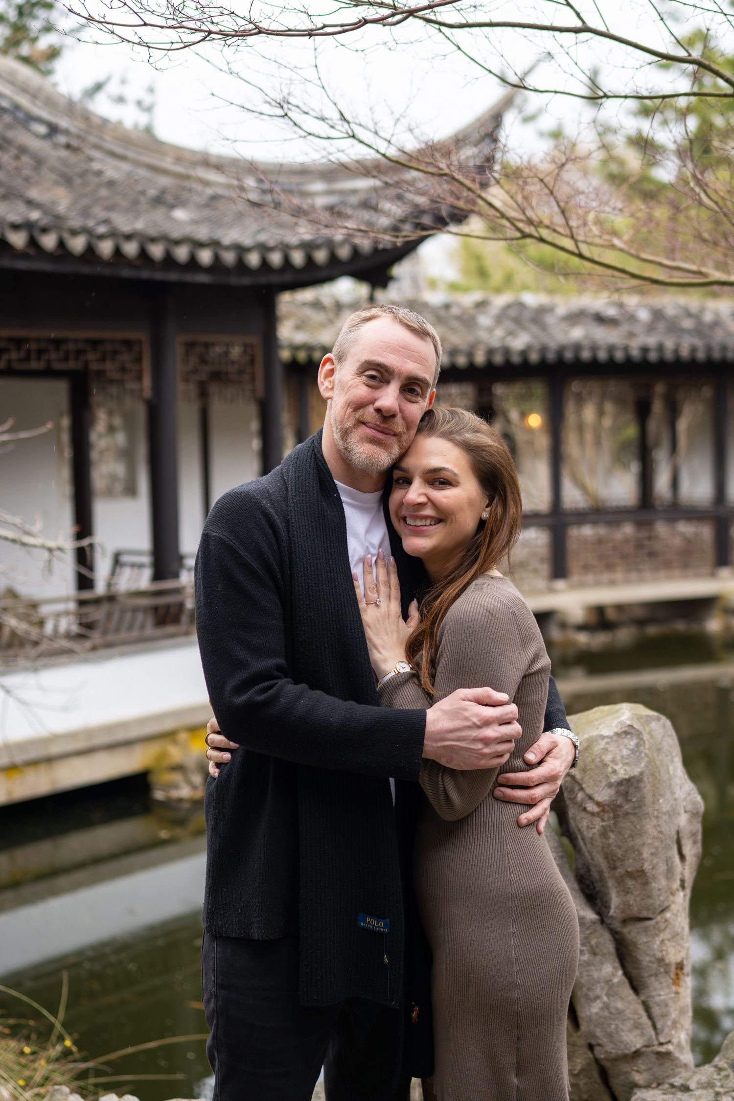 Engaged couple embracing warmly beside a pond with traditional Chinese pavilion in the background.