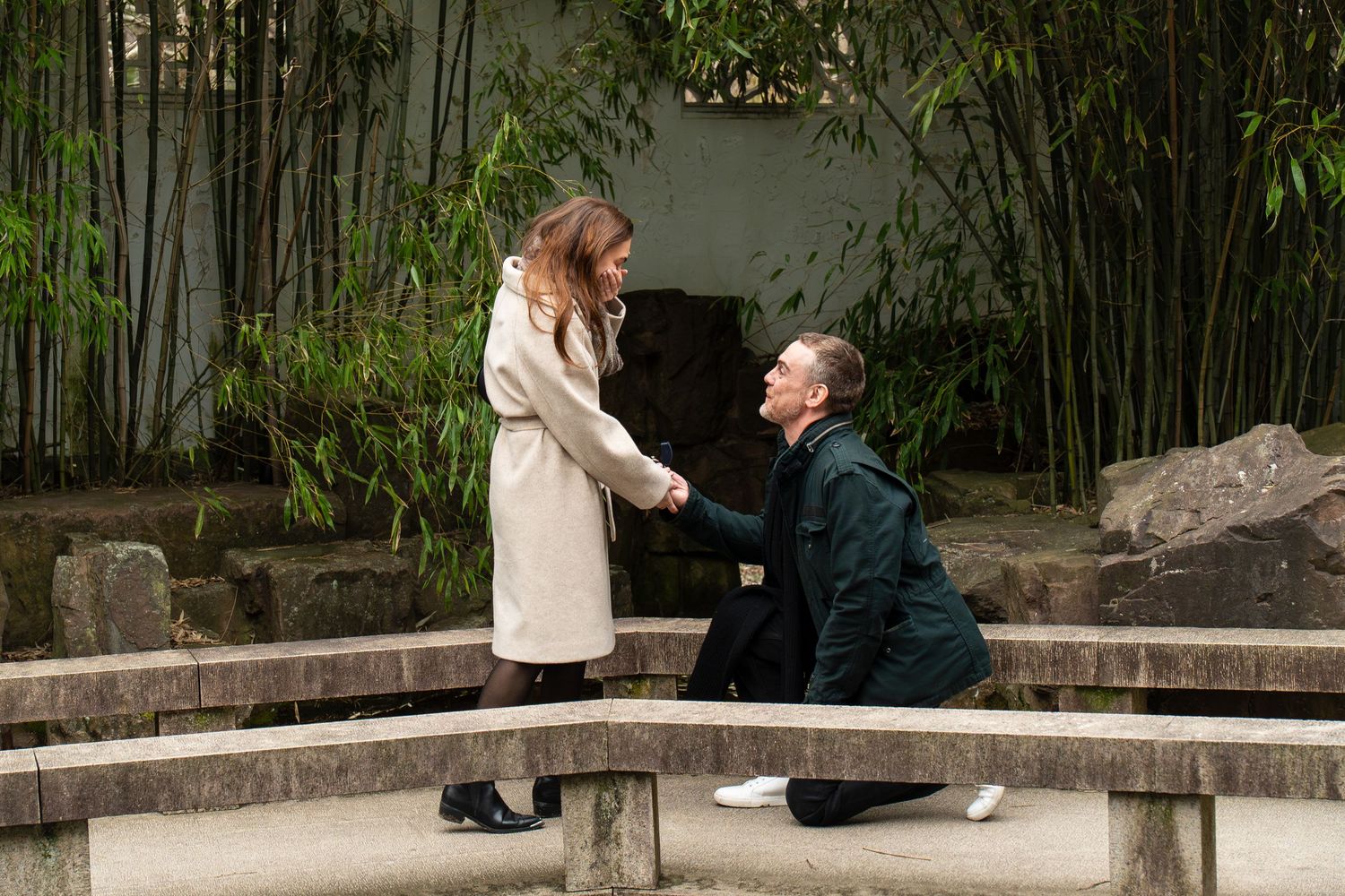 Man kneeling on stone bridge holding woman's hands during romantic marriage proposal in bamboo garden.