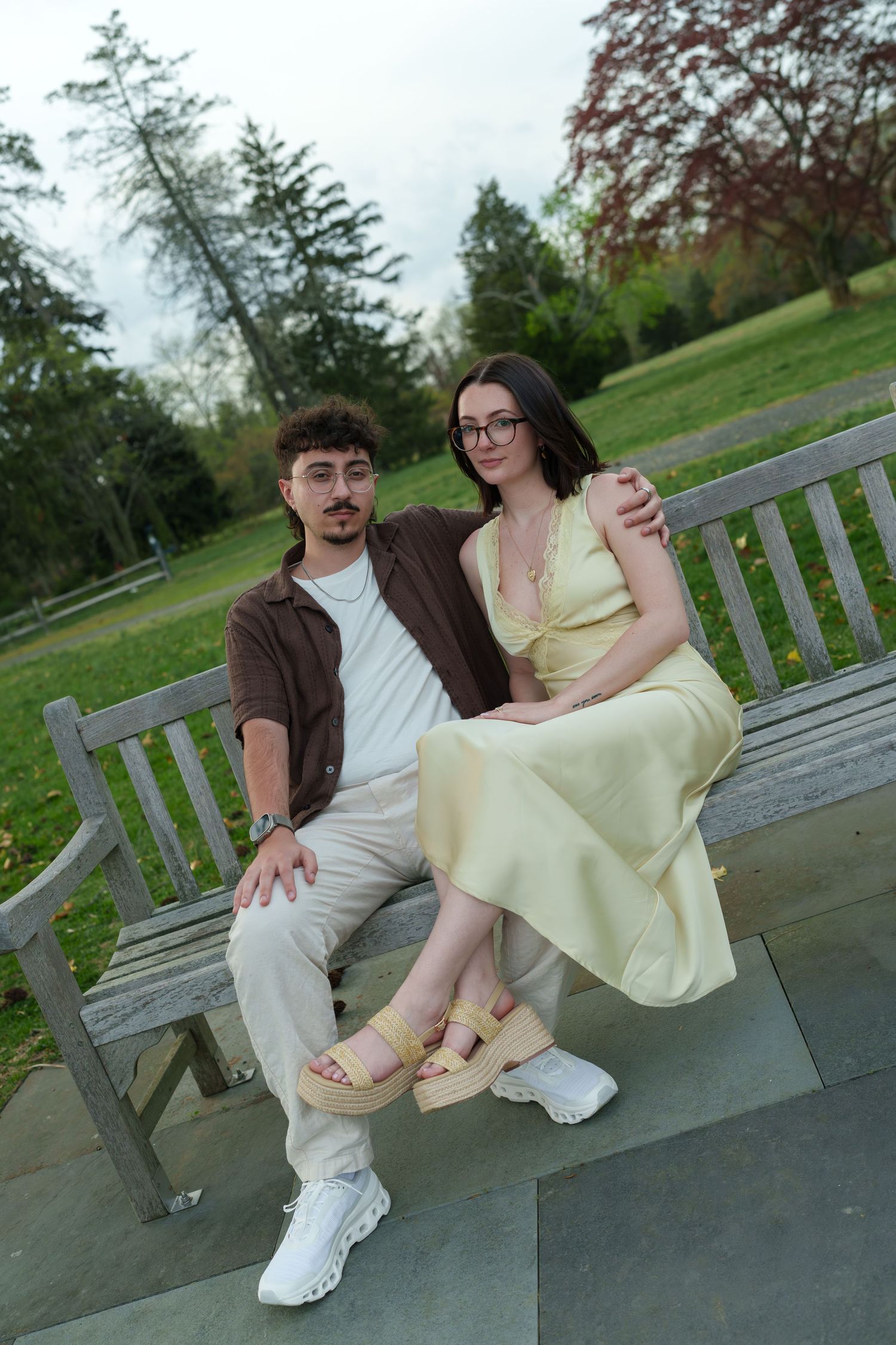 Couple sitting together on a wooden park bench, man in brown shirt and woman in yellow dress.