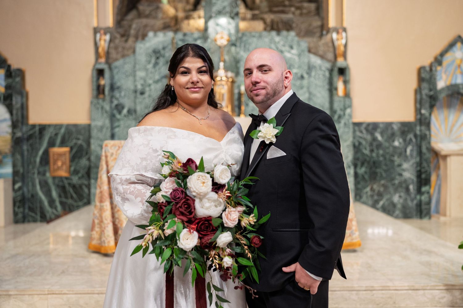 Bride and groom portrait in elegant church with marble backdrop, bride holding burgundy and white bouquet.