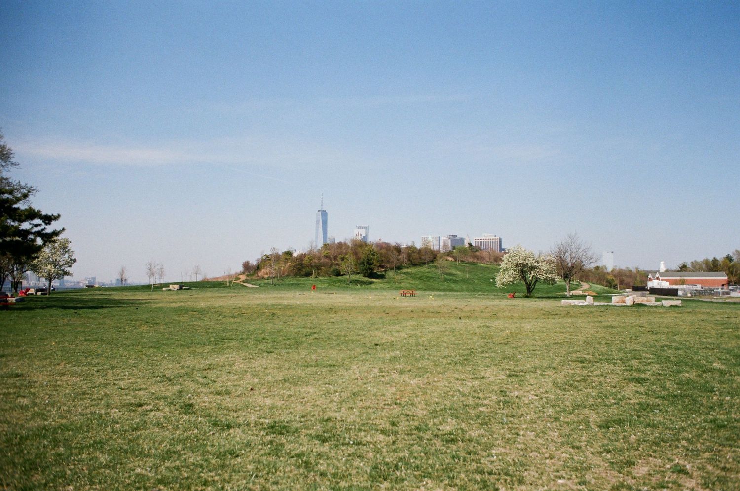 Wide open green grassy field with a hill and city skyline visible in the distance on clear day.
