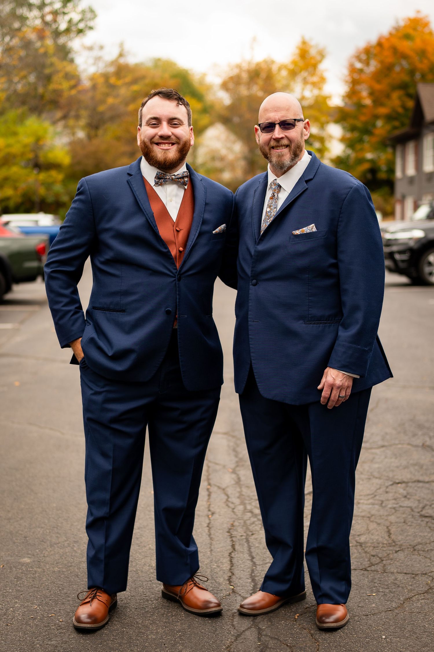 Two men in matching navy suits pose together outdoors among autumn foliage at a fall wedding.