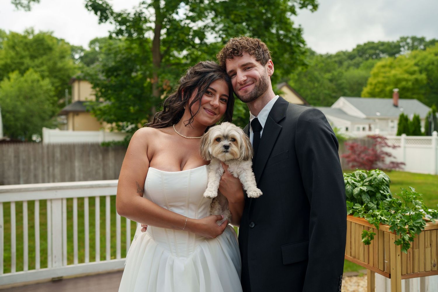 Newlyweds in wedding attire smile holding a small fluffy dog on an outdoor deck.