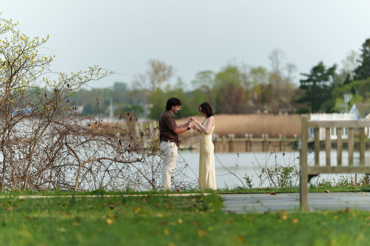 Newly engaged couple holding hands and smiling together on a serene lakeside boardwalk.