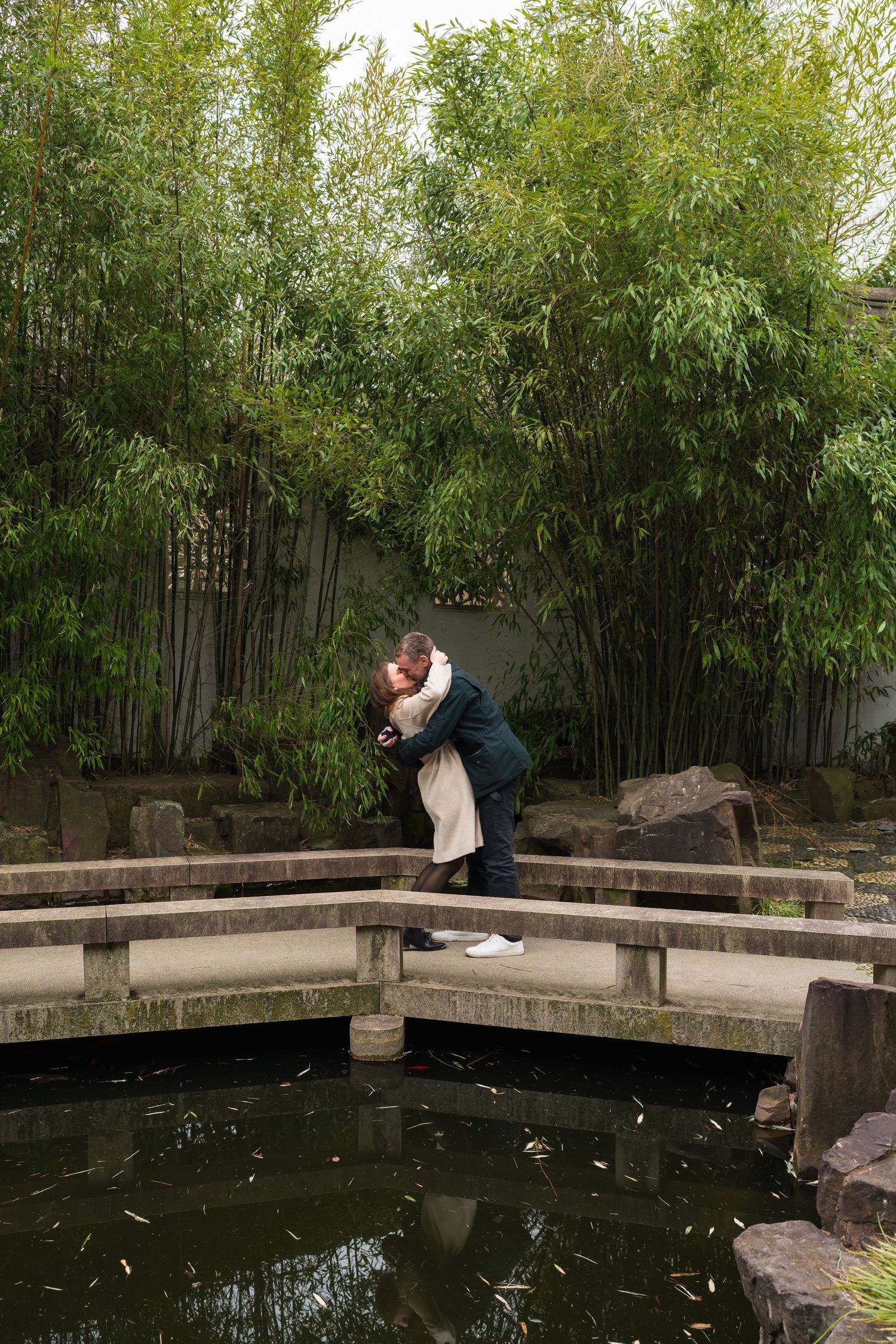 Couple embracing on a stone bridge over a koi pond surrounded by lush bamboo in a Chinese garden.