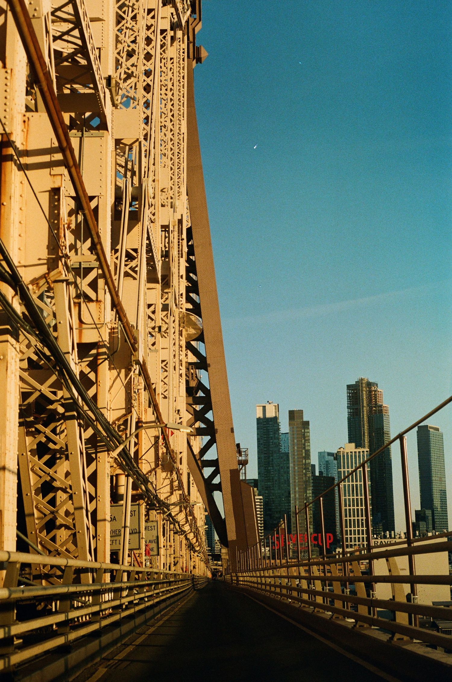 Driving across a massive steel bridge with NYC skyline skyscrapers visible under a clear blue sky.