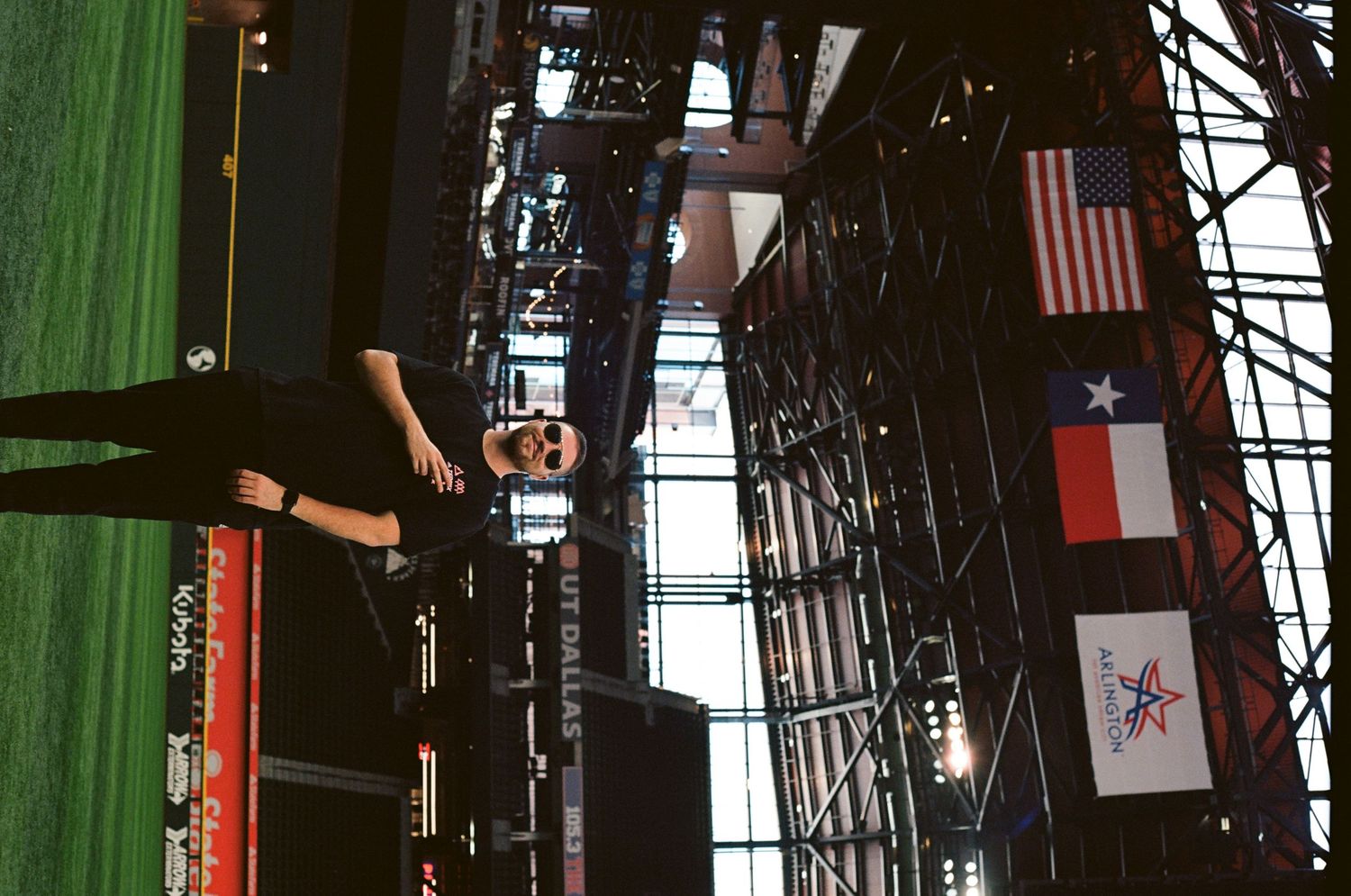 Person standing inside a stadium with American and Texas flags visible on metal scaffolding above a green field.