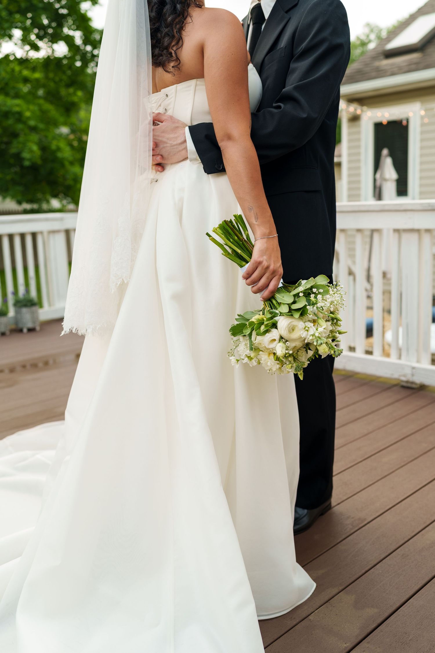 Bride and groom embracing on a wooden deck, bride holding a white and green wedding bouquet in an elegant white gown.