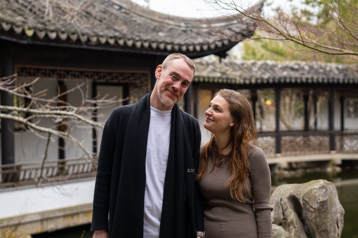 Couple smiling together in front of a traditional Chinese garden pavilion with a tranquil pond.