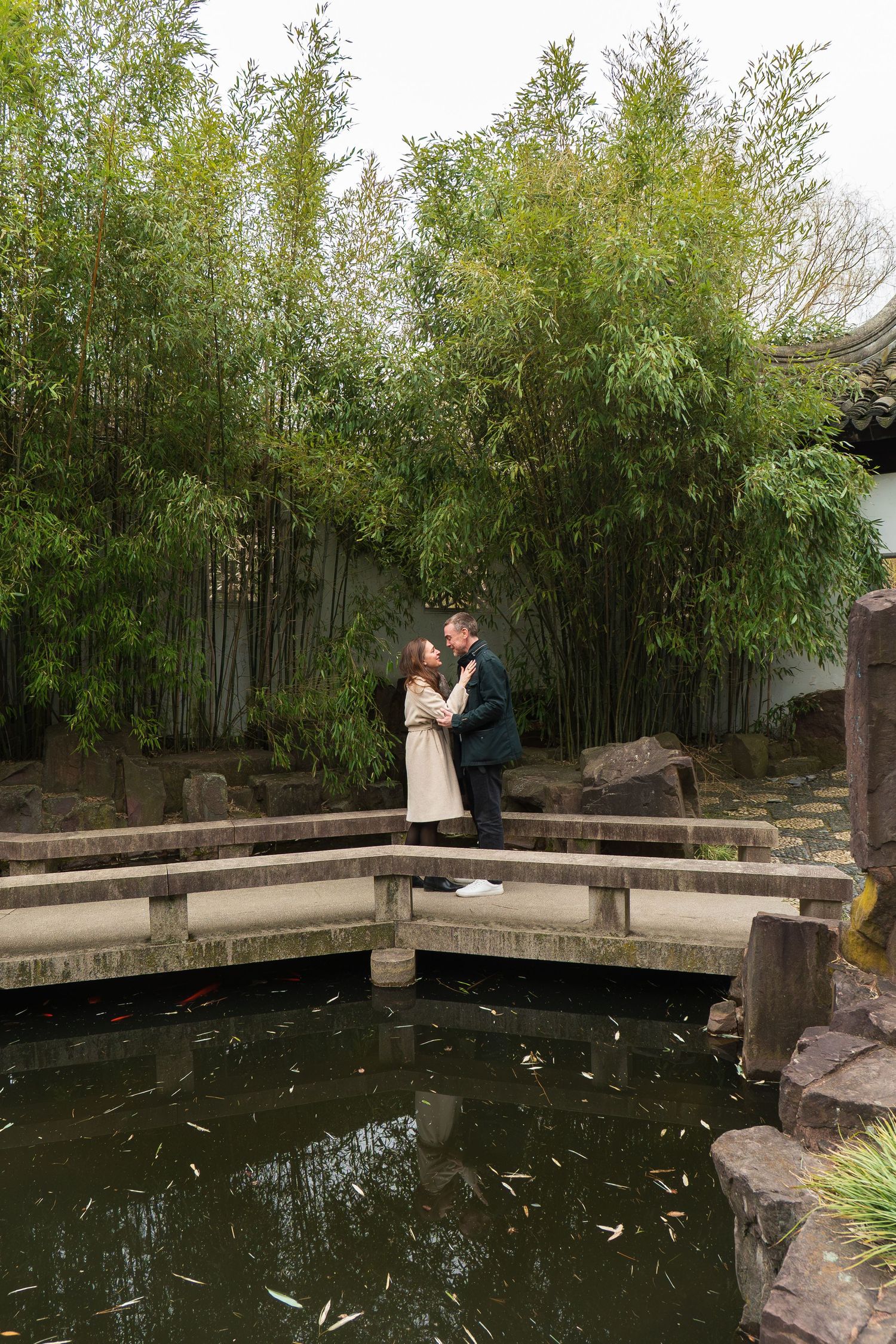 Couple embracing and kissing on a stone bridge over a koi pond surrounded by tall bamboo trees.