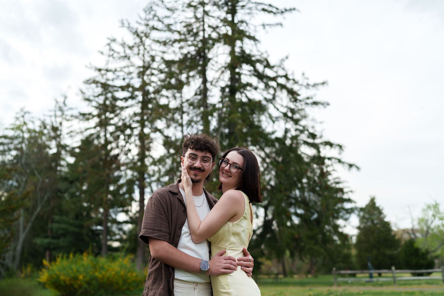 Couple embracing and smiling in a lush green park with tall evergreen trees in the background.