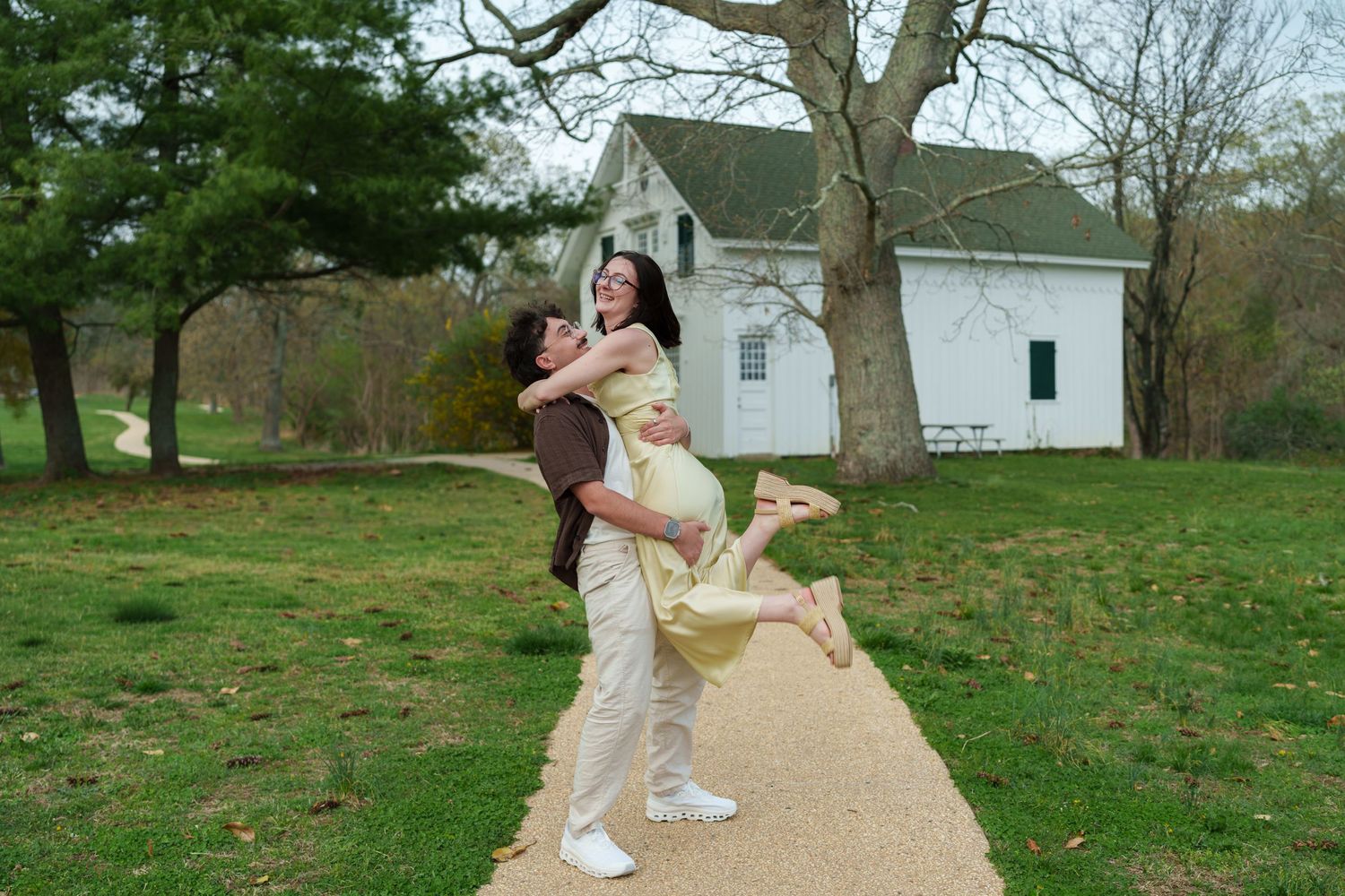 Man joyfully lifting woman in yellow dress on a garden path near a white countryside building.