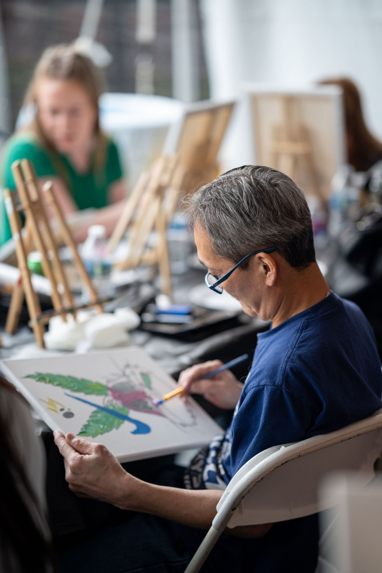 Man with glasses painting colorful artwork on canvas at an indoor art workshop.
