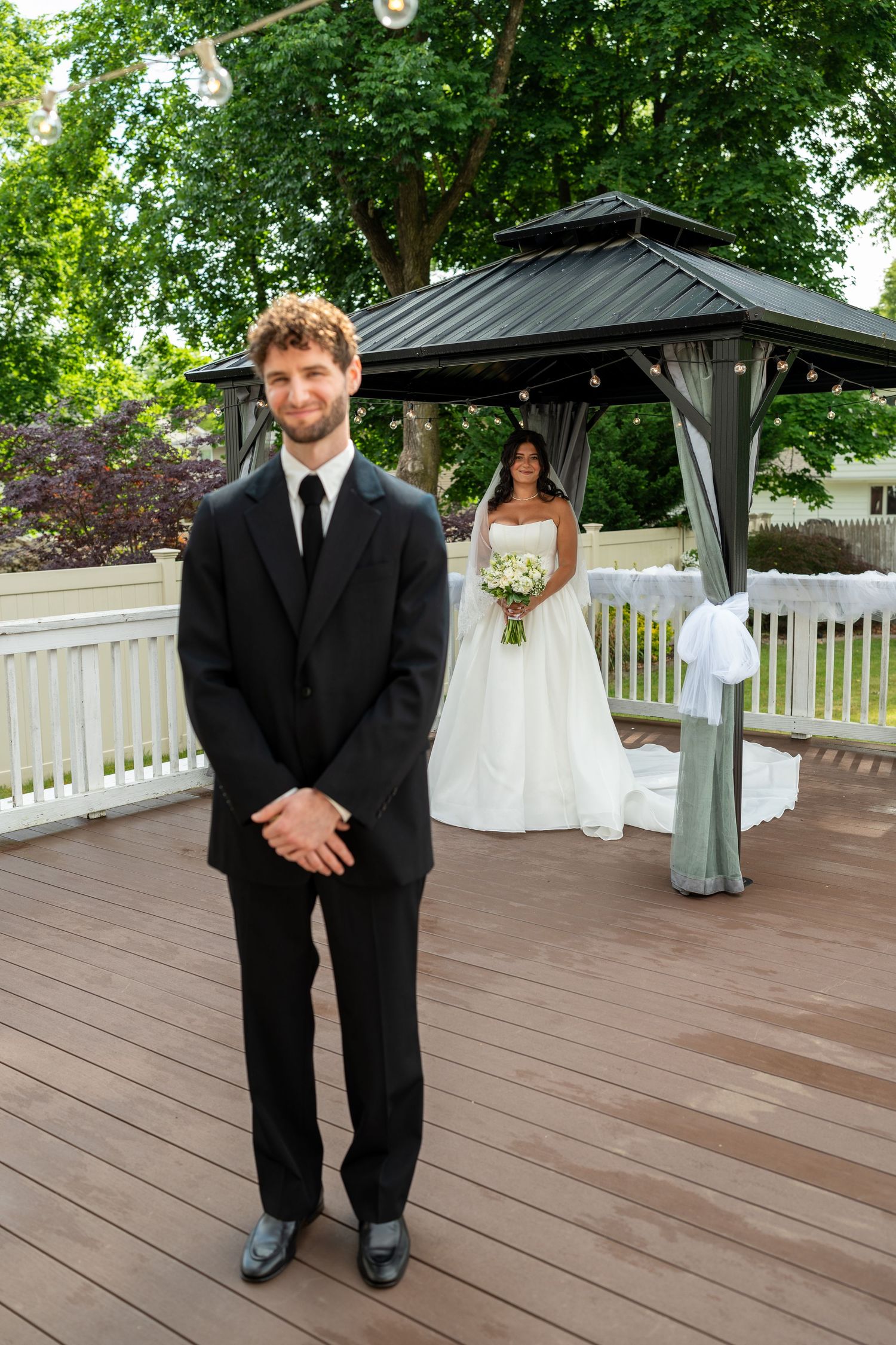 Groom in black suit stands on deck as bride in white gown approaches during first look.