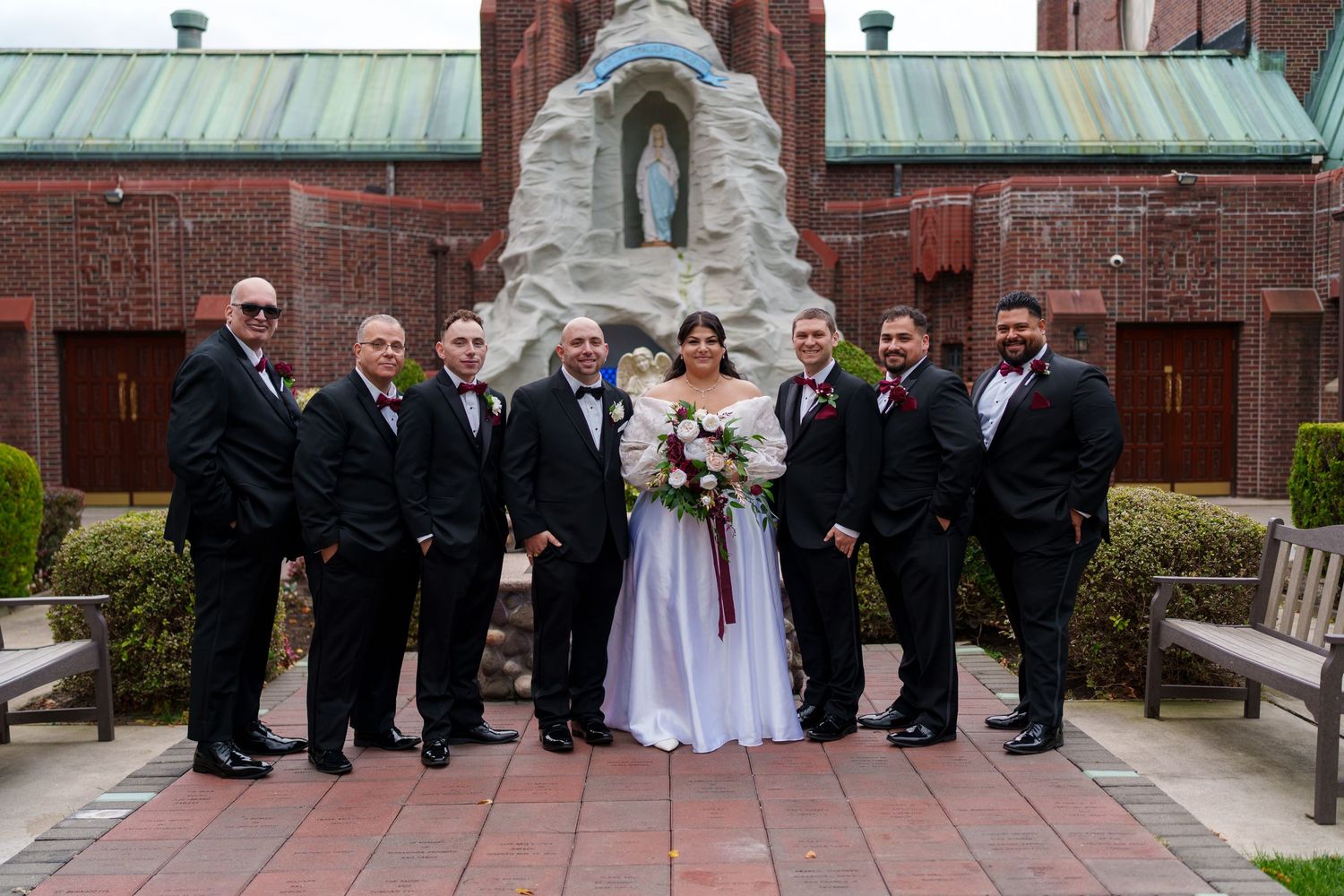 Wedding party in black suits posing with bride in white gown before a church grotto.