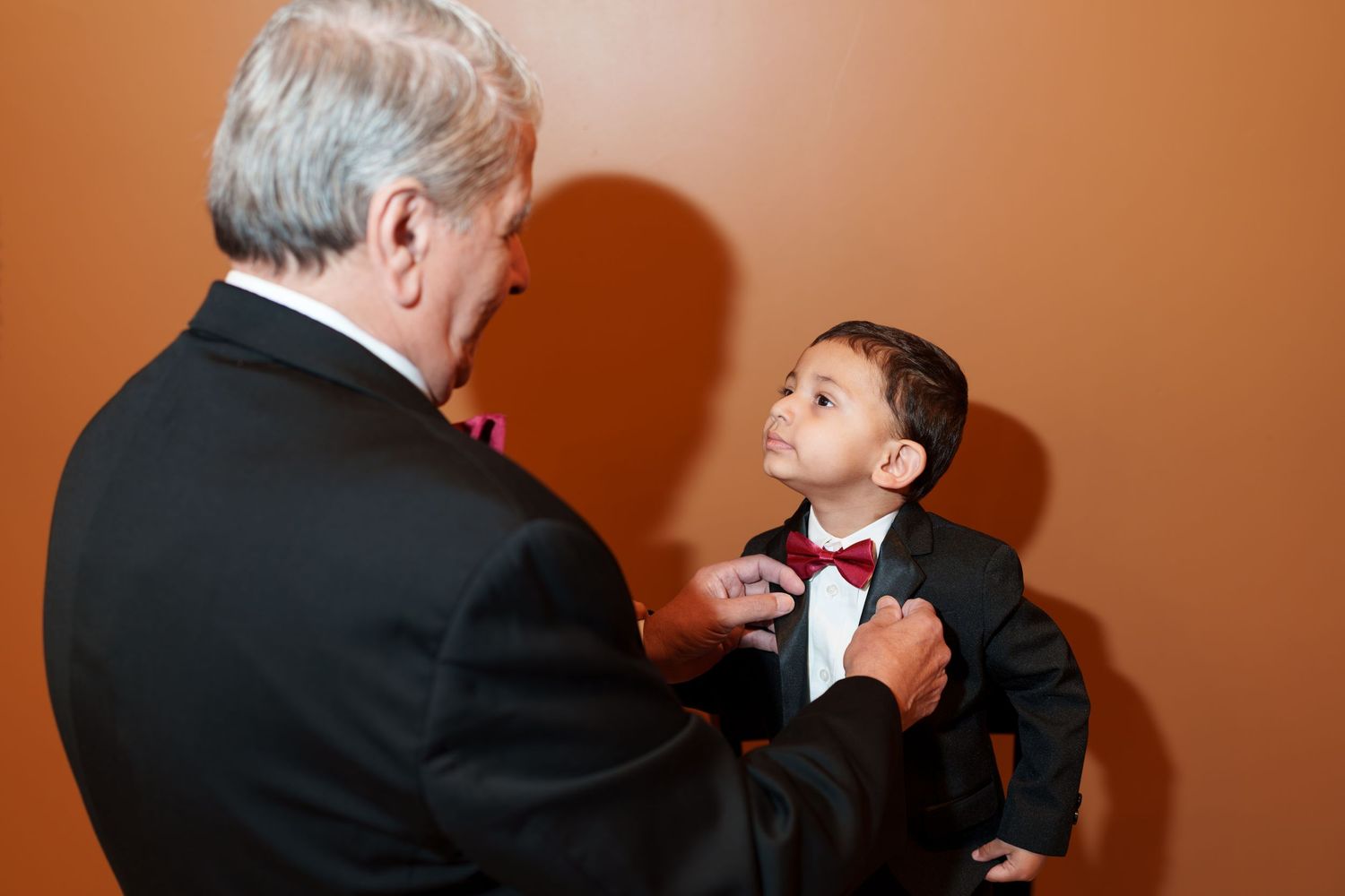 Older man in tuxedo adjusting bow tie of young boy dressed in formal black suit.