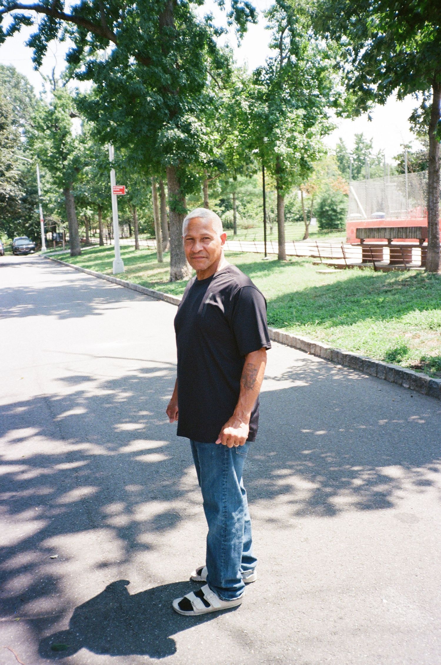 A man in a black t-shirt and jeans stands on a tree-lined suburban street on a sunny day.