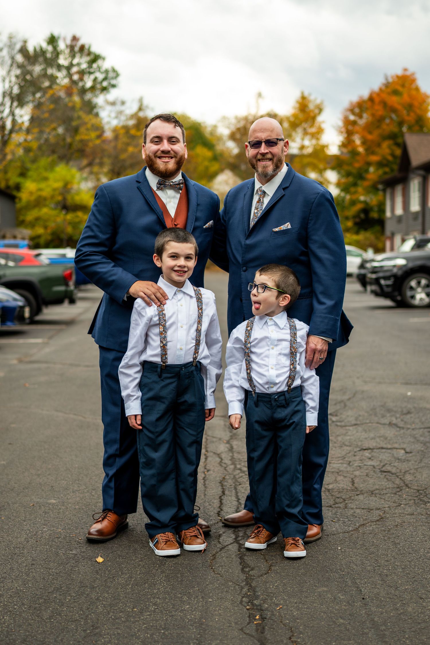 Two men in navy suits pose proudly with two young boys in suspenders at fall wedding.
