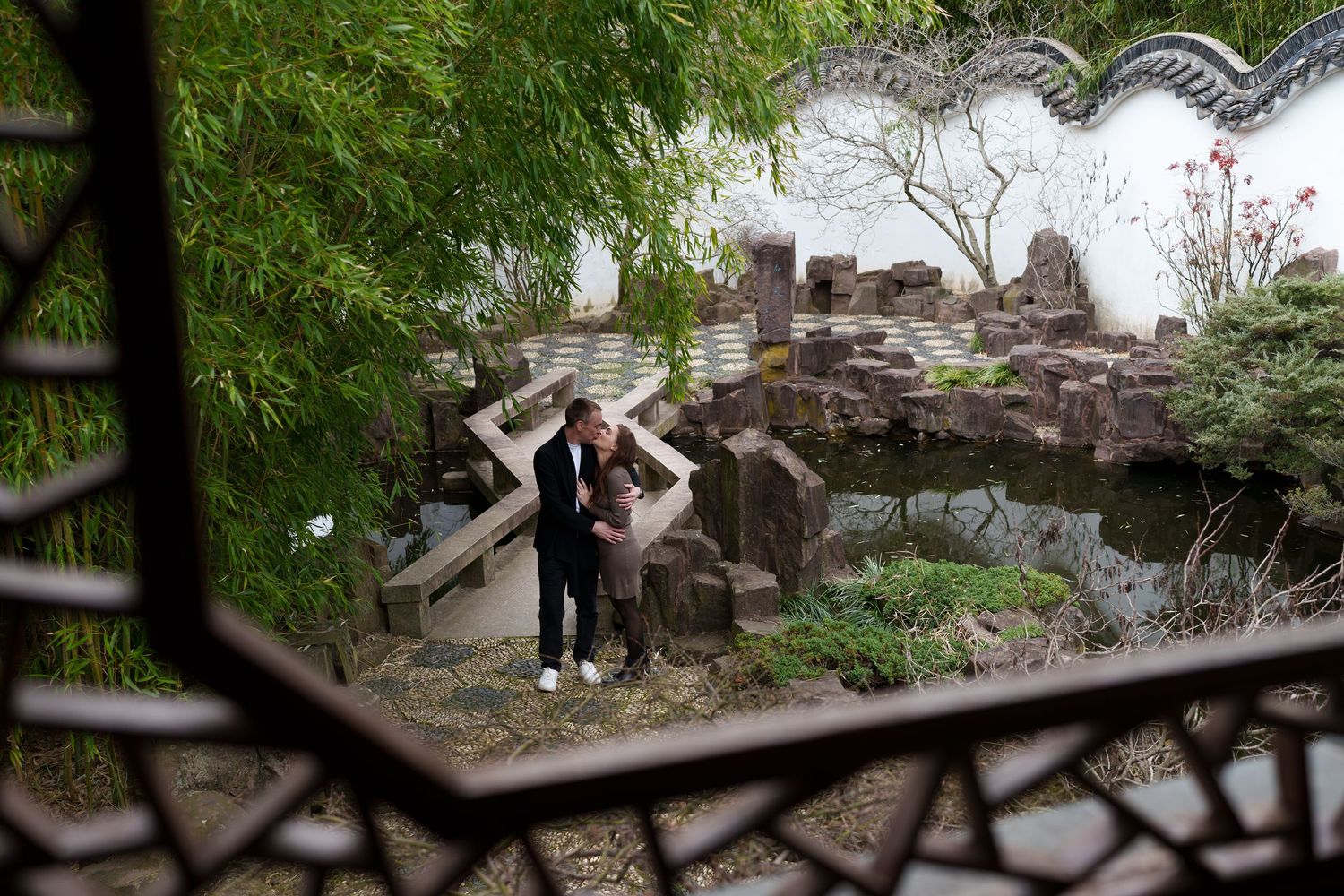 Couple embracing near a koi pond and rockery viewed through an ornate lattice window in a Chinese garden.