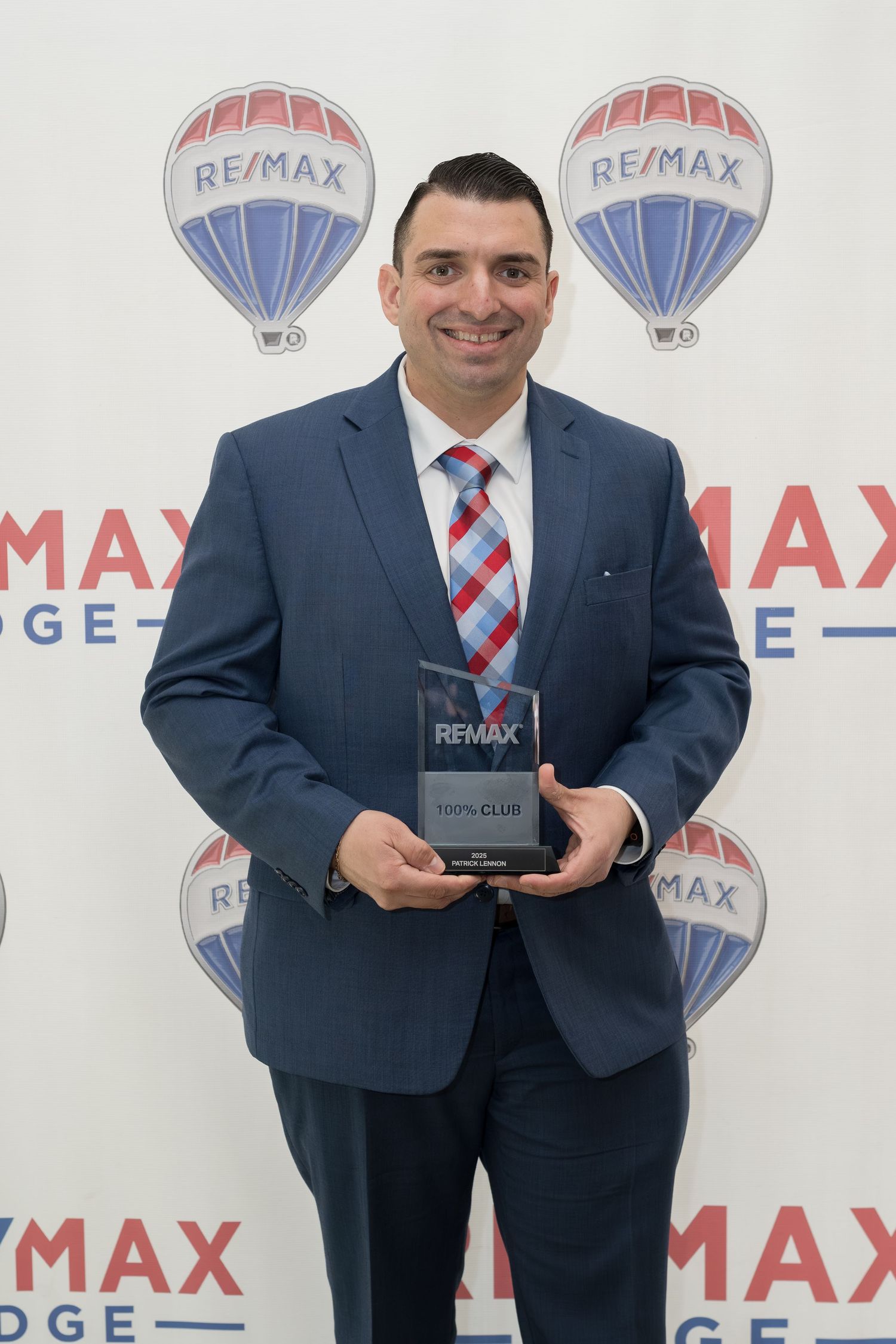 Man in blue suit displays RE/MAX 100% Club award at ceremony.