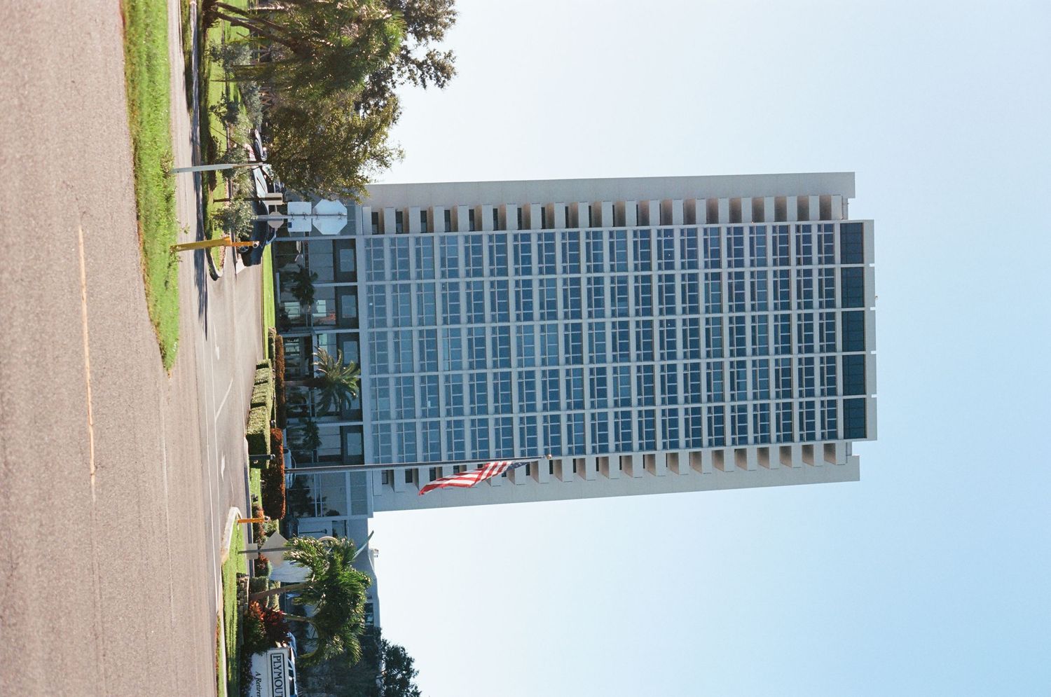 Tall modern office building with glass facade surrounded by trees under a clear blue sky.