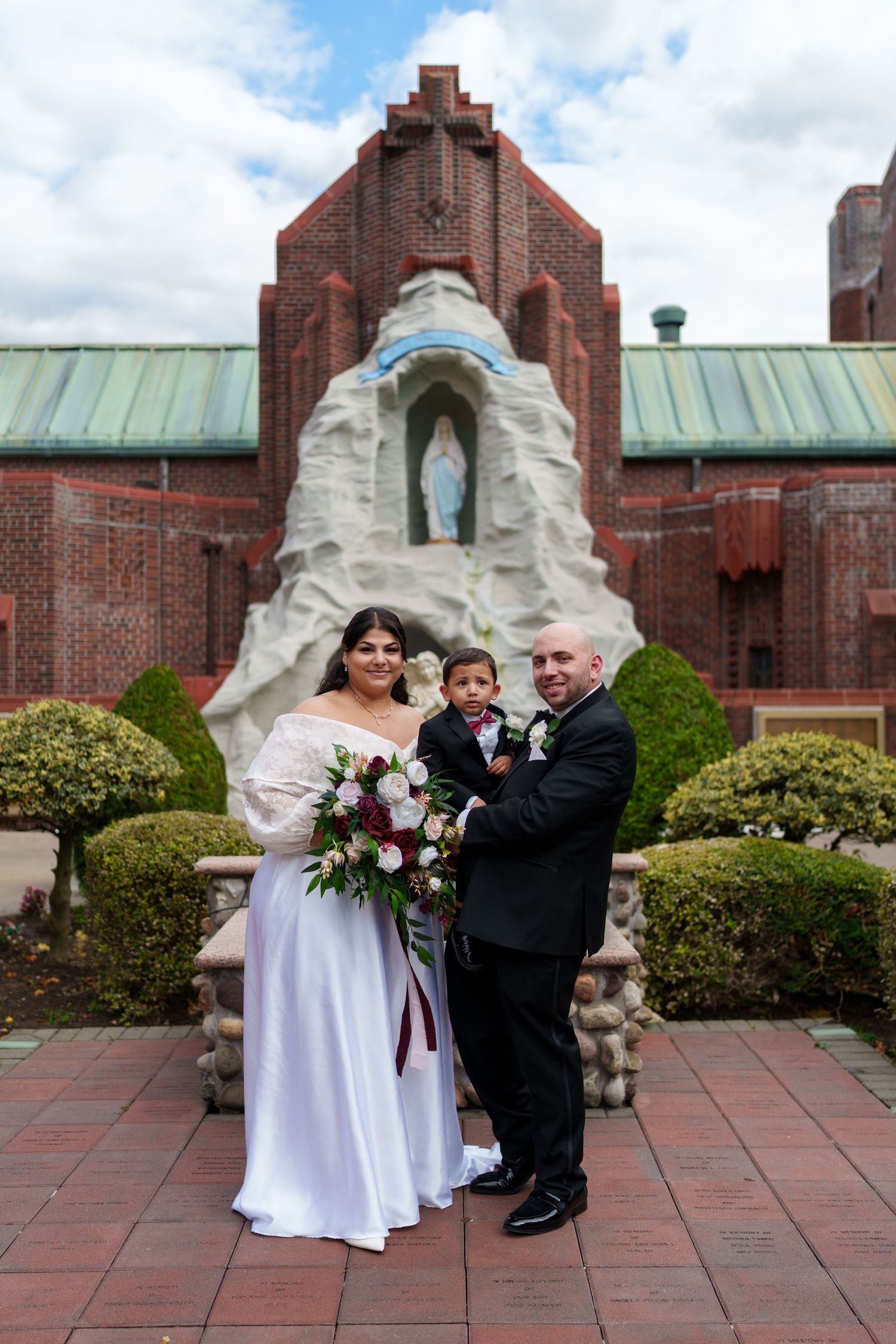 Bride, groom, and young son pose before church grotto statue with lush white and burgundy bouquet.