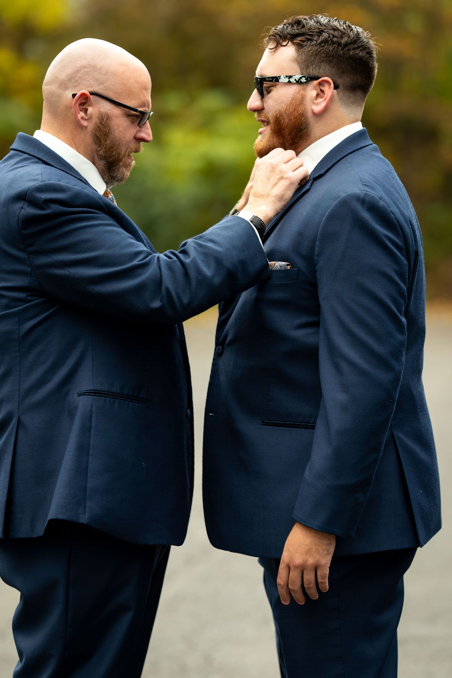 Two groomsmen in navy suits adjust each other's attire outdoors at a fall wedding.