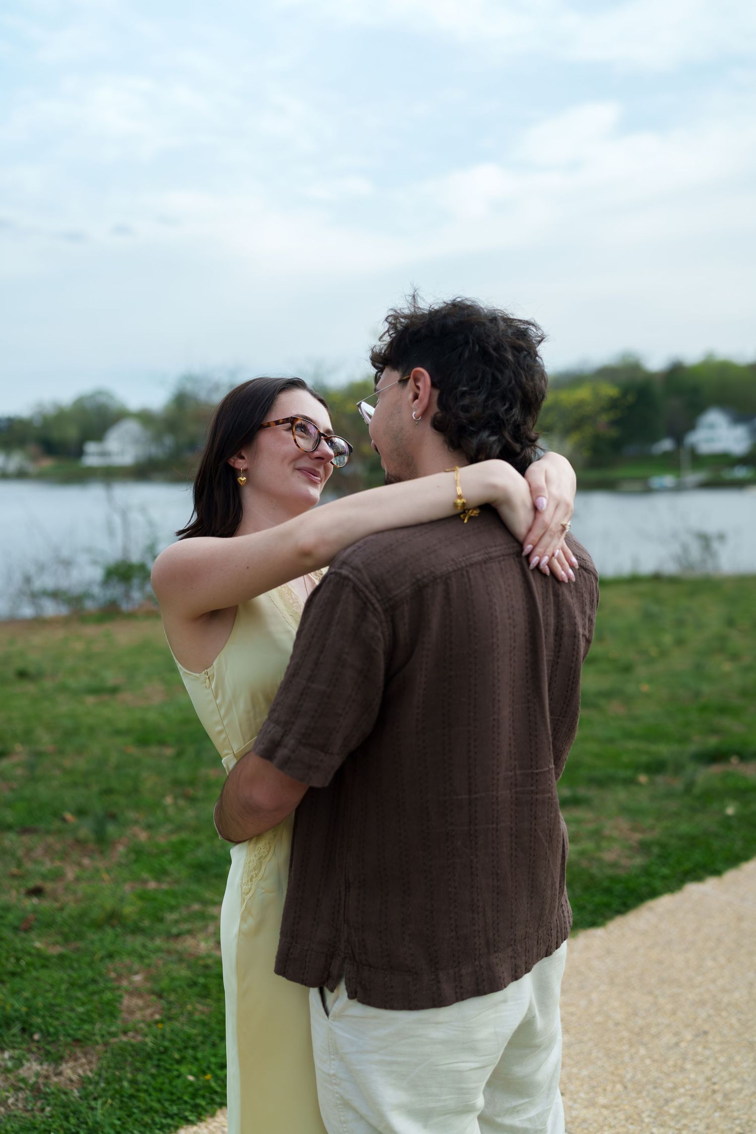 Couple embracing and smiling at each other near a lake in a park, woman in yellow dress and man in brown shirt.