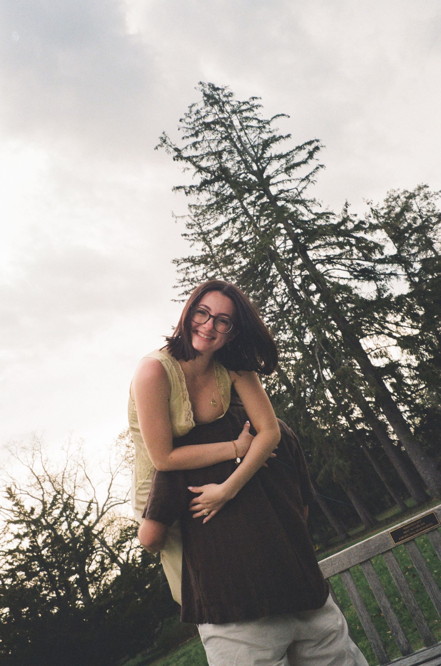 Laughing woman in a yellow dress embracing her partner outdoors near tall evergreen trees.